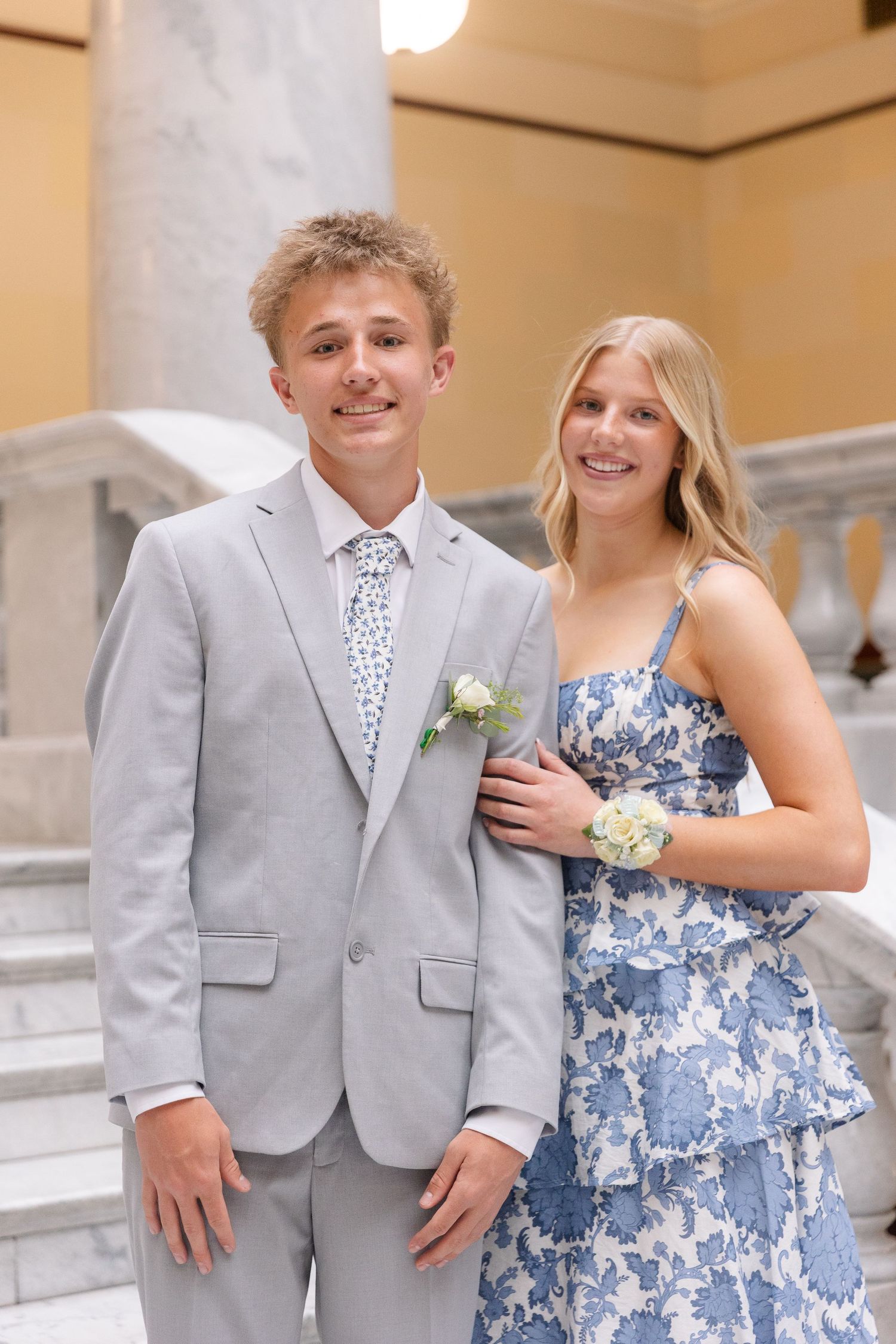 Smiling prom couple posing indoors, boy in gray suit with floral tie and girl in blue floral dress.