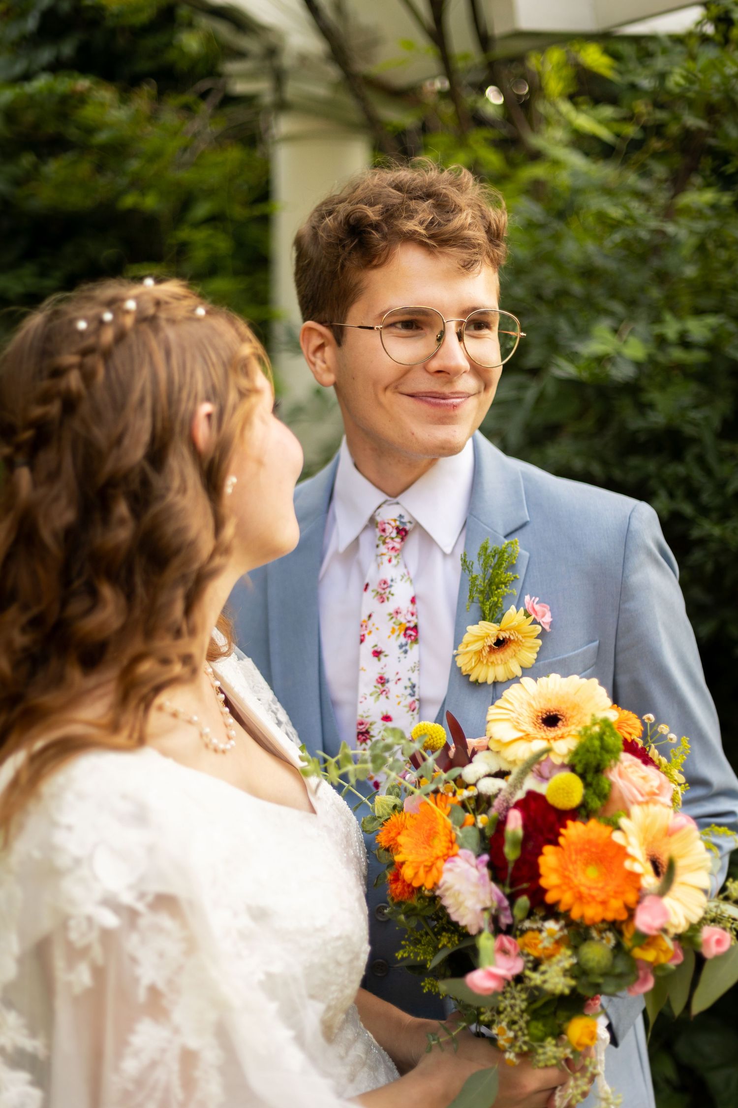 Groom in blue suit with floral tie smiling at bride holding colorful bouquet outdoors.