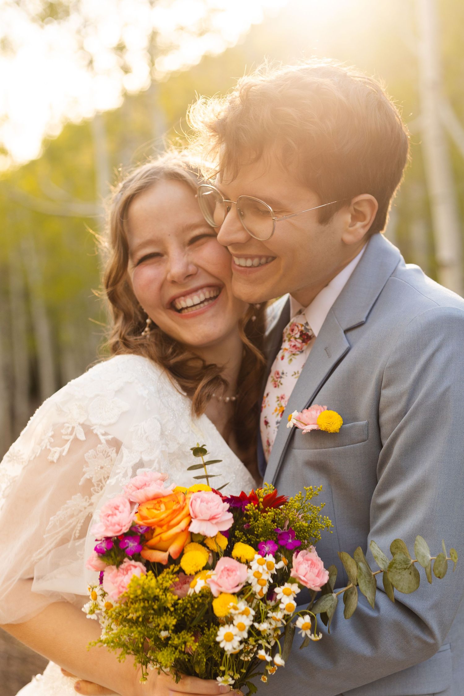 Joyful bride and groom laughing together outdoors, holding colorful wildflower bouquet in golden sunlight.