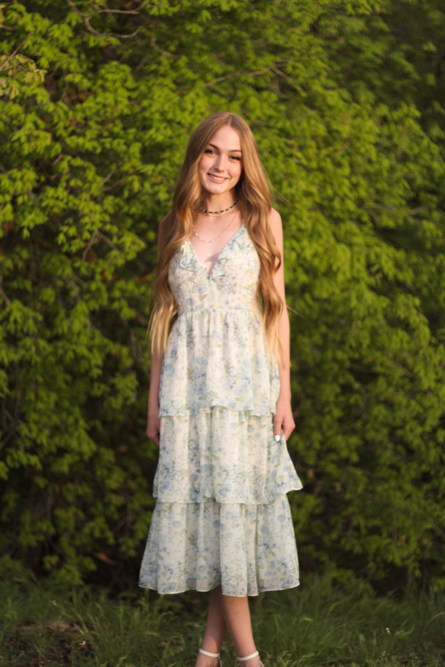 Young woman in tiered floral sundress with long blonde hair smiling in front of lush green trees.