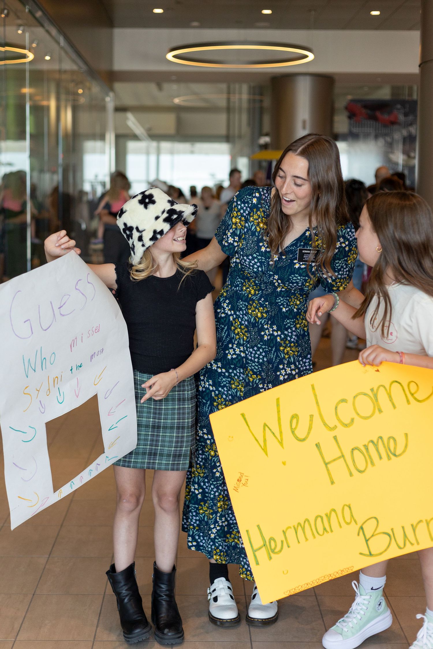 Young woman in floral dress welcomed home by girls holding handmade signs indoors.
