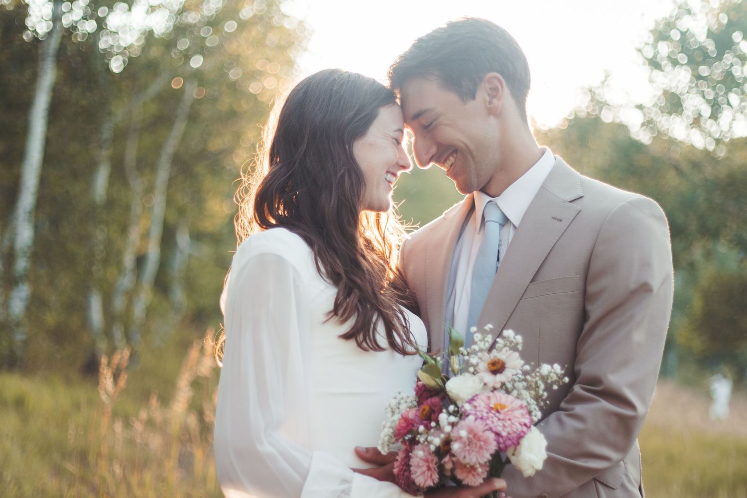 Smiling bride and groom touch foreheads in golden sunlight among birch trees, bride holding pink floral bouquet.