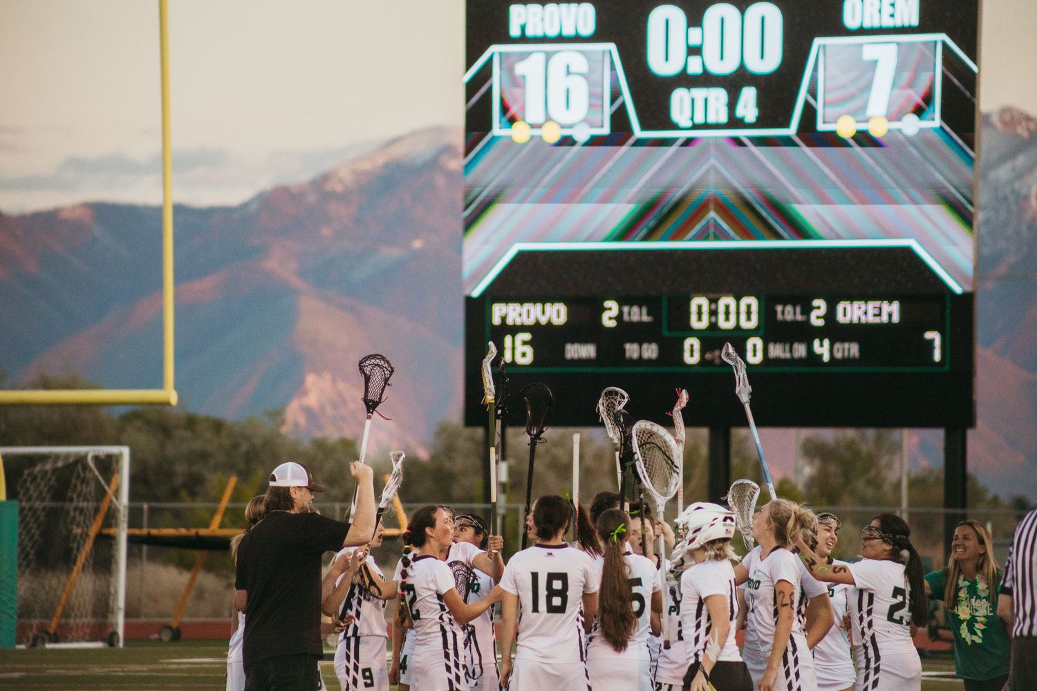 Lacrosse team celebrates a 16-7 victory over Orem at end of 4th quarter under a scoreboard with mountains behind.