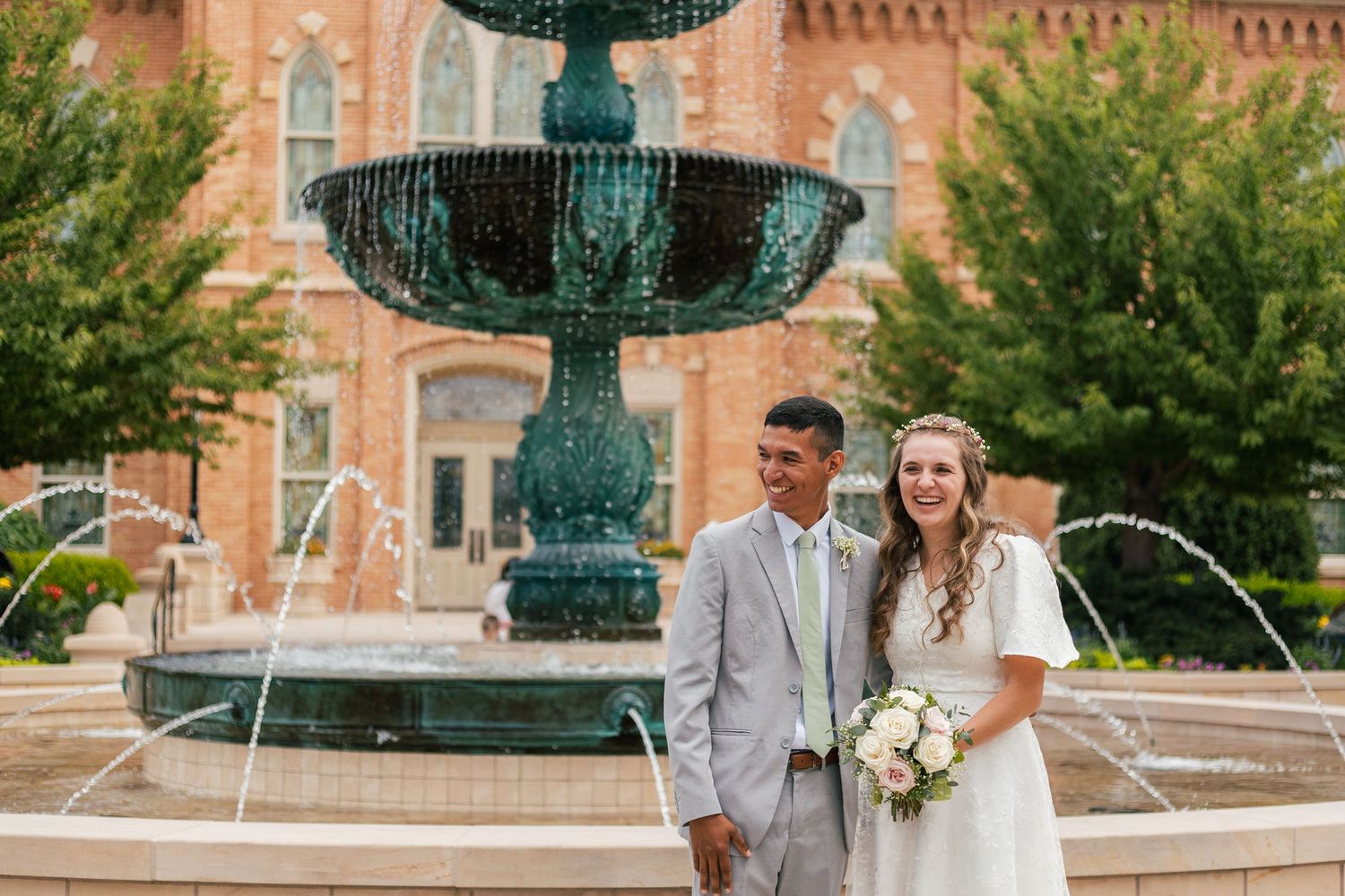 Bride and groom smiling together in front of a green tiered fountain outside a brick building.