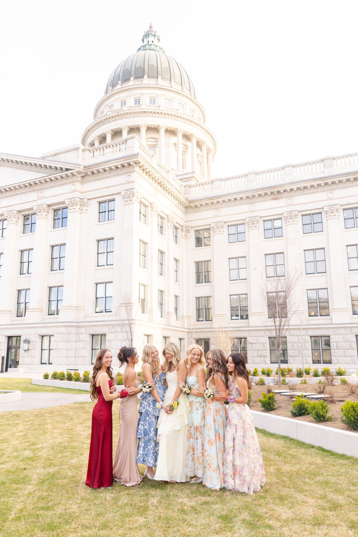 Six young women in formal prom dresses posing in front of a large domed capitol building.
