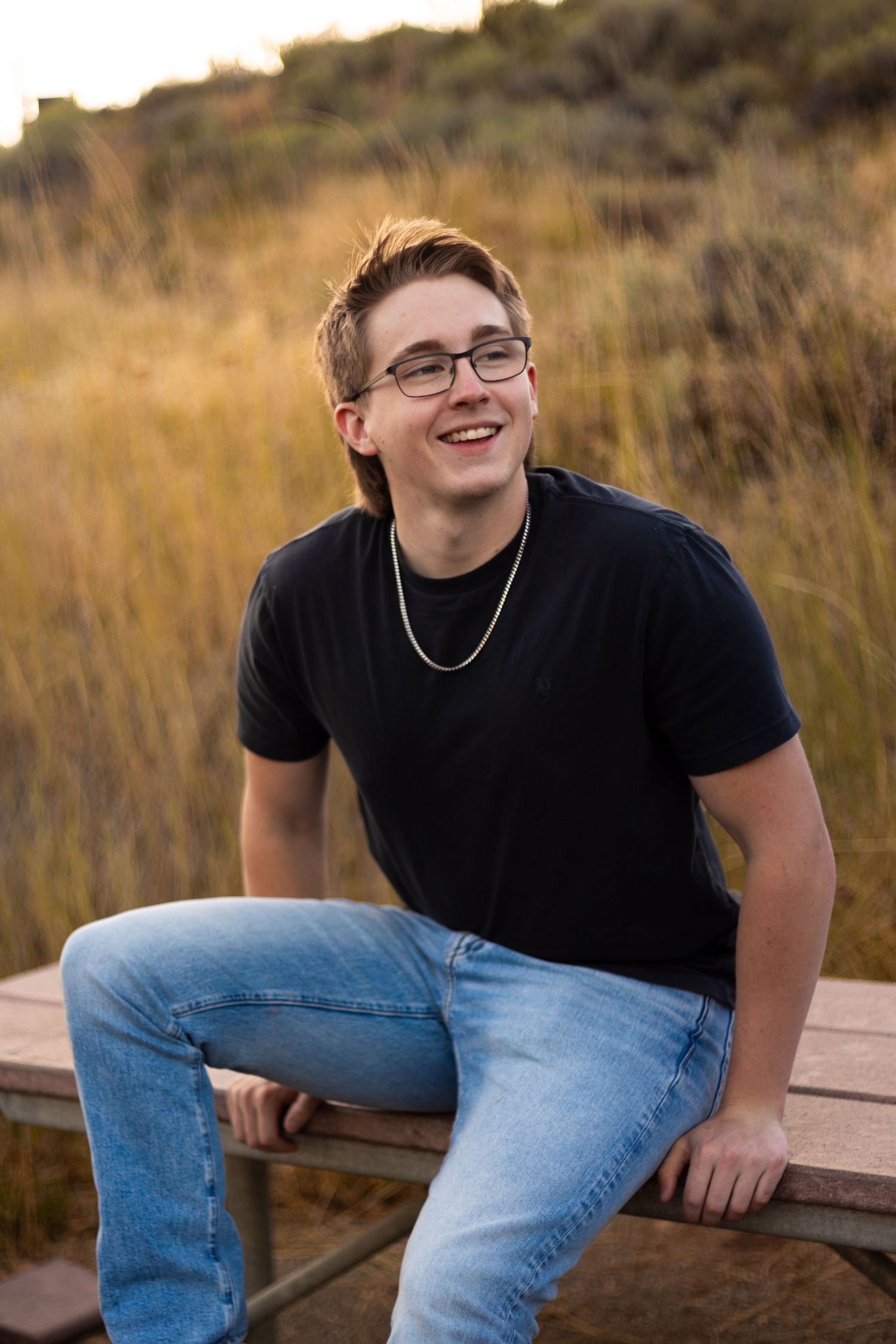 Teenage boy with glasses in black t-shirt sitting on wooden bench in golden field.