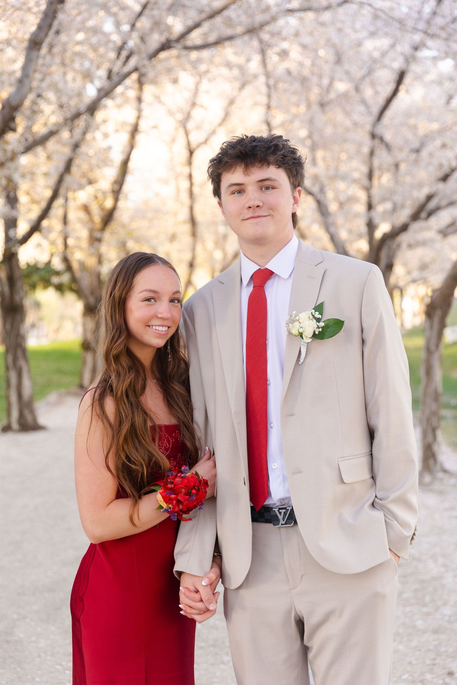 Prom couple holding hands among cherry blossoms, girl in red dress and boy in tan suit with red tie.