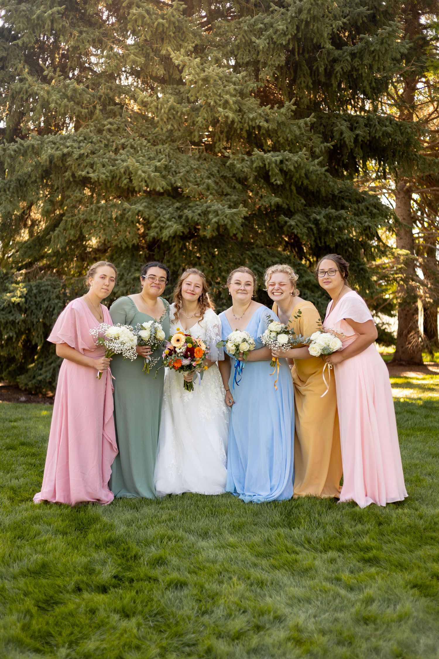 Bride with five bridesmaids in colorful mismatched gowns holding bouquets outdoors near tall evergreen trees.