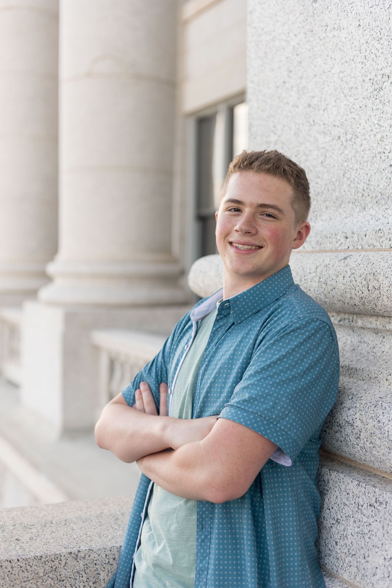 Smiling teenage boy in a teal button-up shirt leaning against a stone column with arms crossed outdoors.