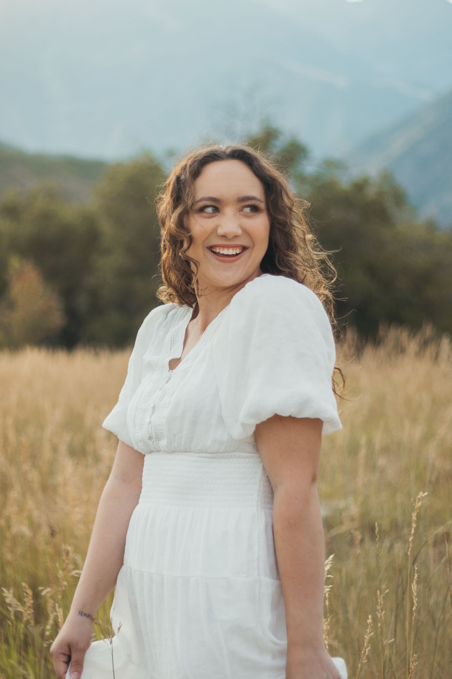 Curly-haired woman in white dress laughs joyfully in golden grass field with misty mountains behind her.