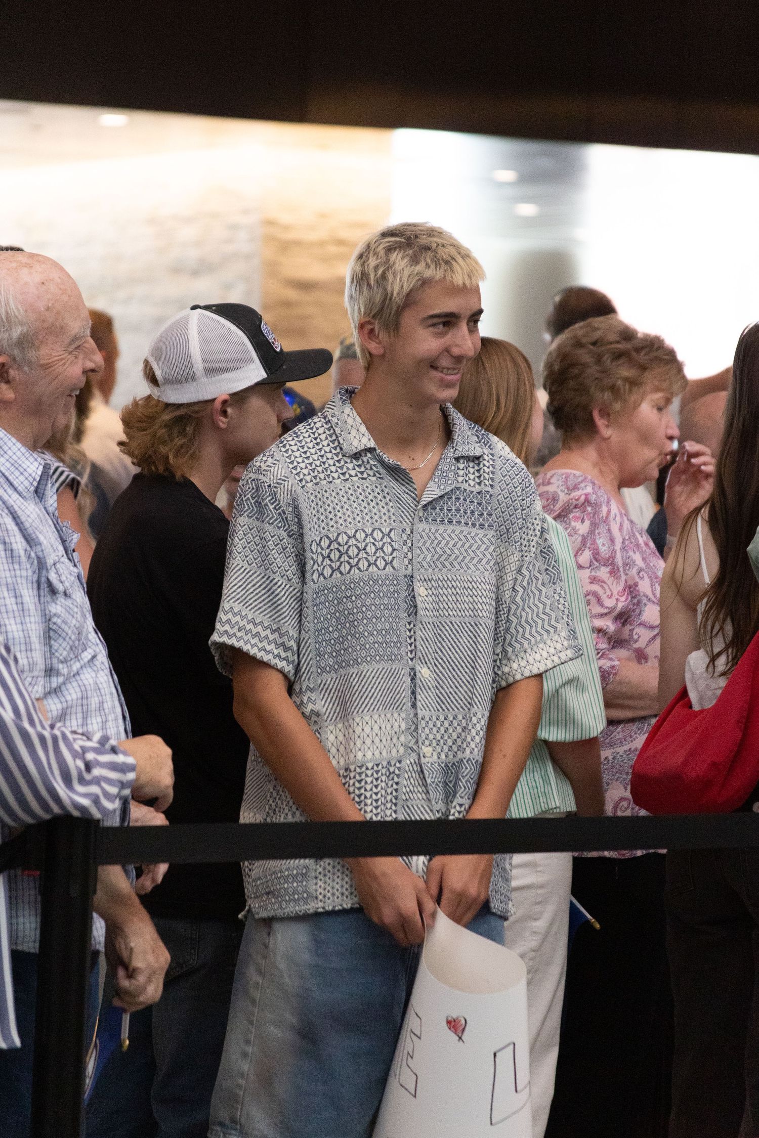 Smiling young man with blonde hair wearing a patterned shirt stands in an airport arrival queue for his sister to return from her mission.