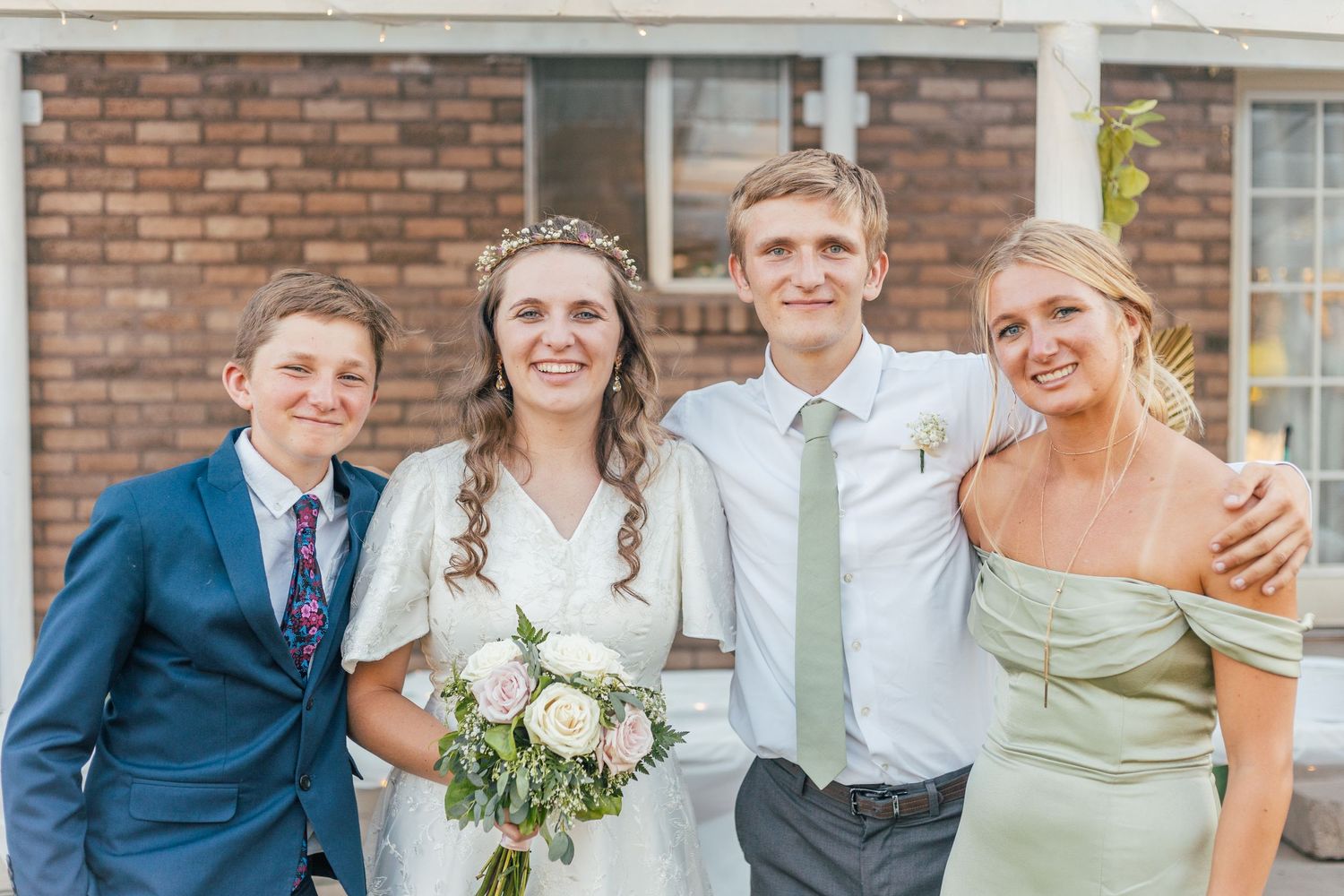 Wedding party of four posing outdoors, bride in white gown holding bouquet with groom and two siblings.