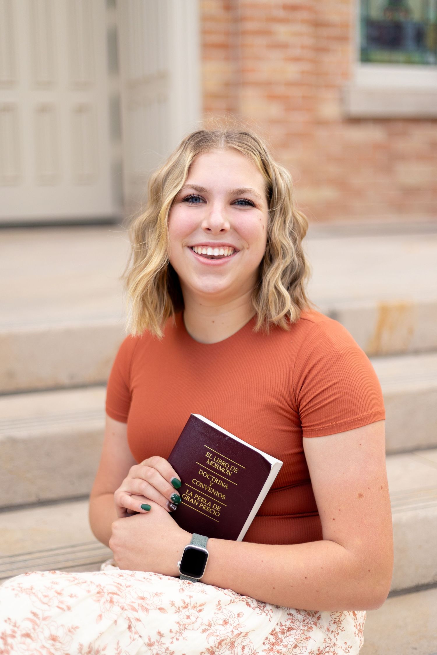 Smiling young blonde woman in an orange top holding a book, sitting on steps near a brick building outdoors.