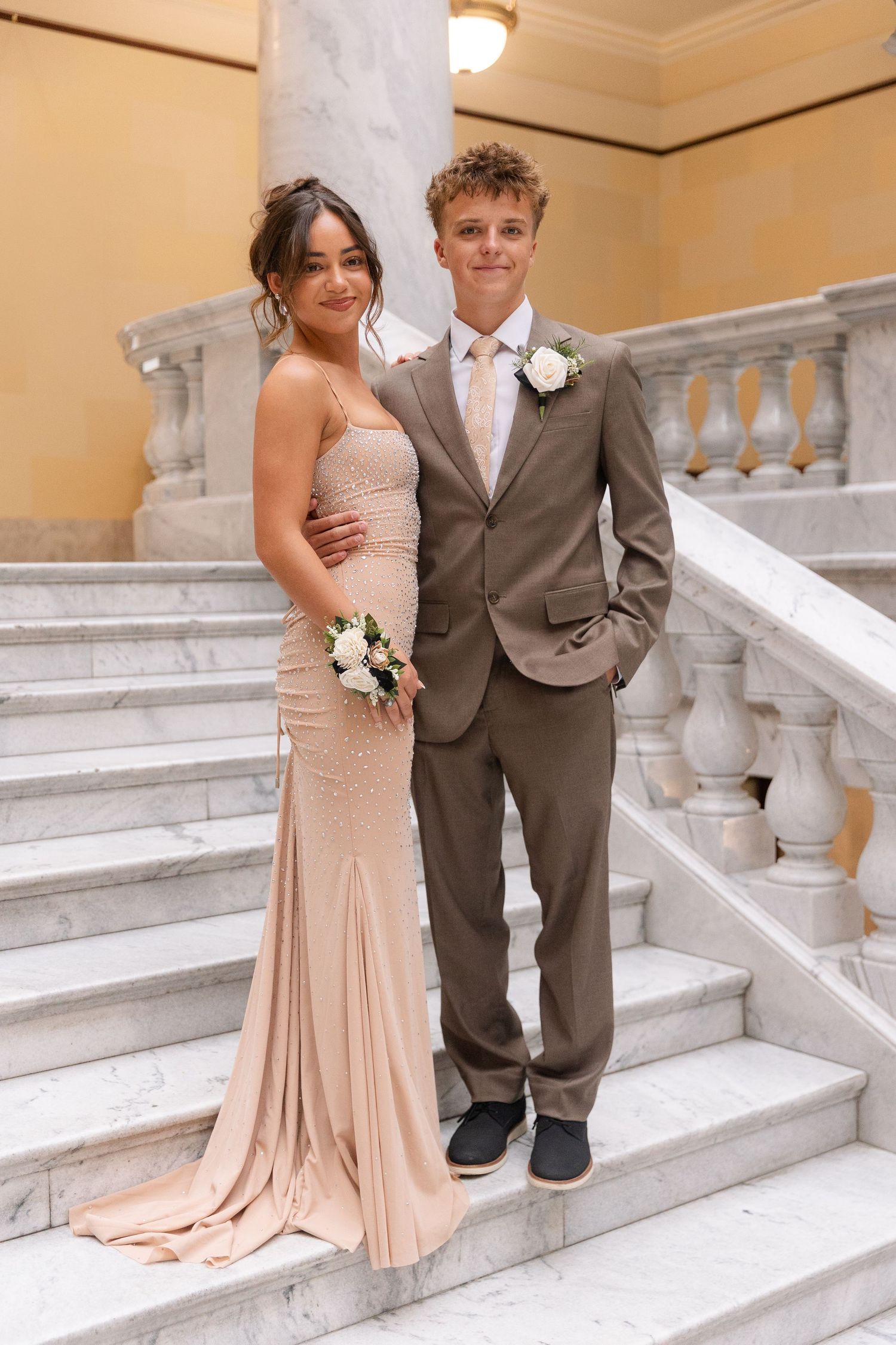Prom couple on marble staircase, girl in champagne beaded gown and boy in taupe suit with pink tie.