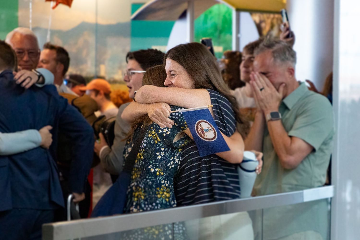 Mom and return missionary daughter share a joyful embrace at an airport reunion while holding a small flag.