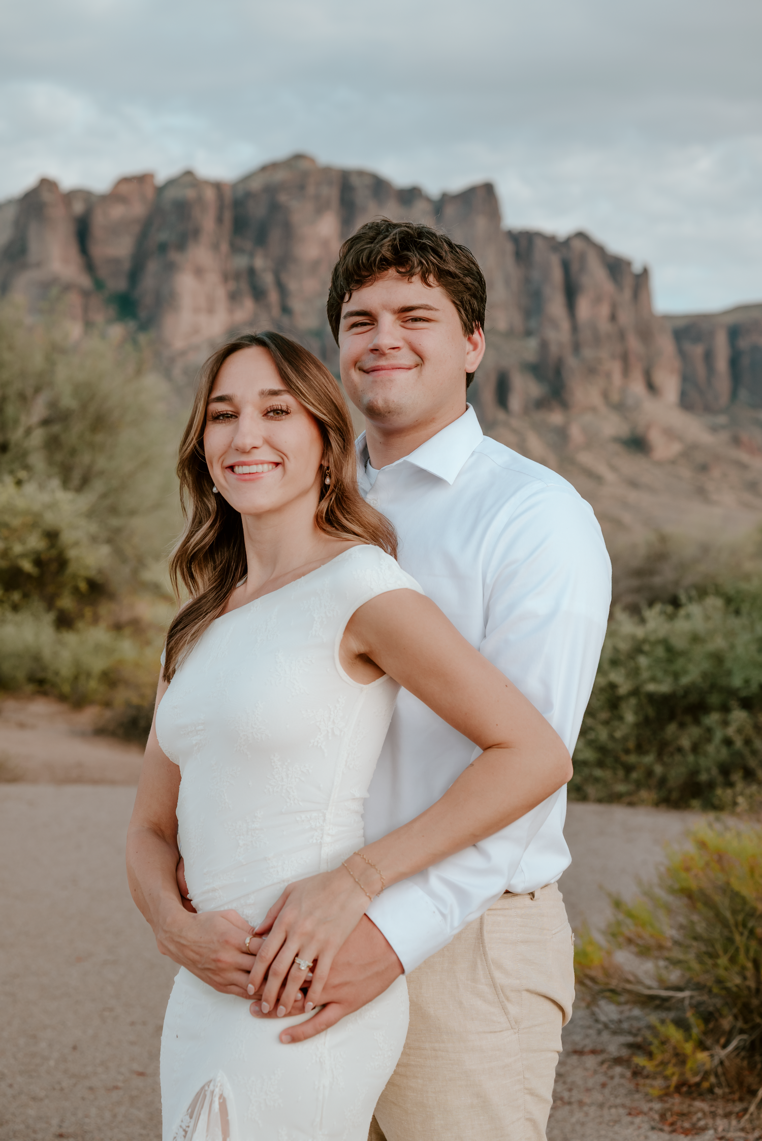 Couple poses for maternity photos in front of Superstition Mountains in Arizona, woman in white dress.