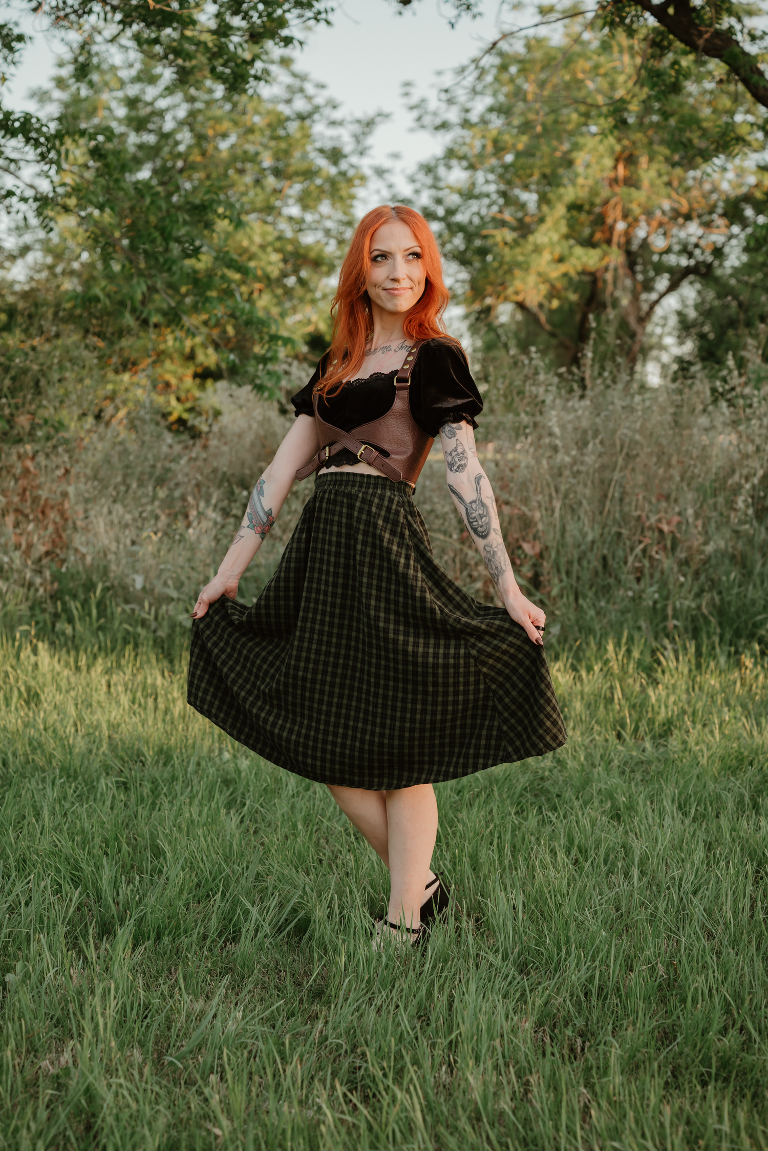Red-haired woman with tattoos wearing a plaid midi skirt and black top posing outdoors in a green meadow.