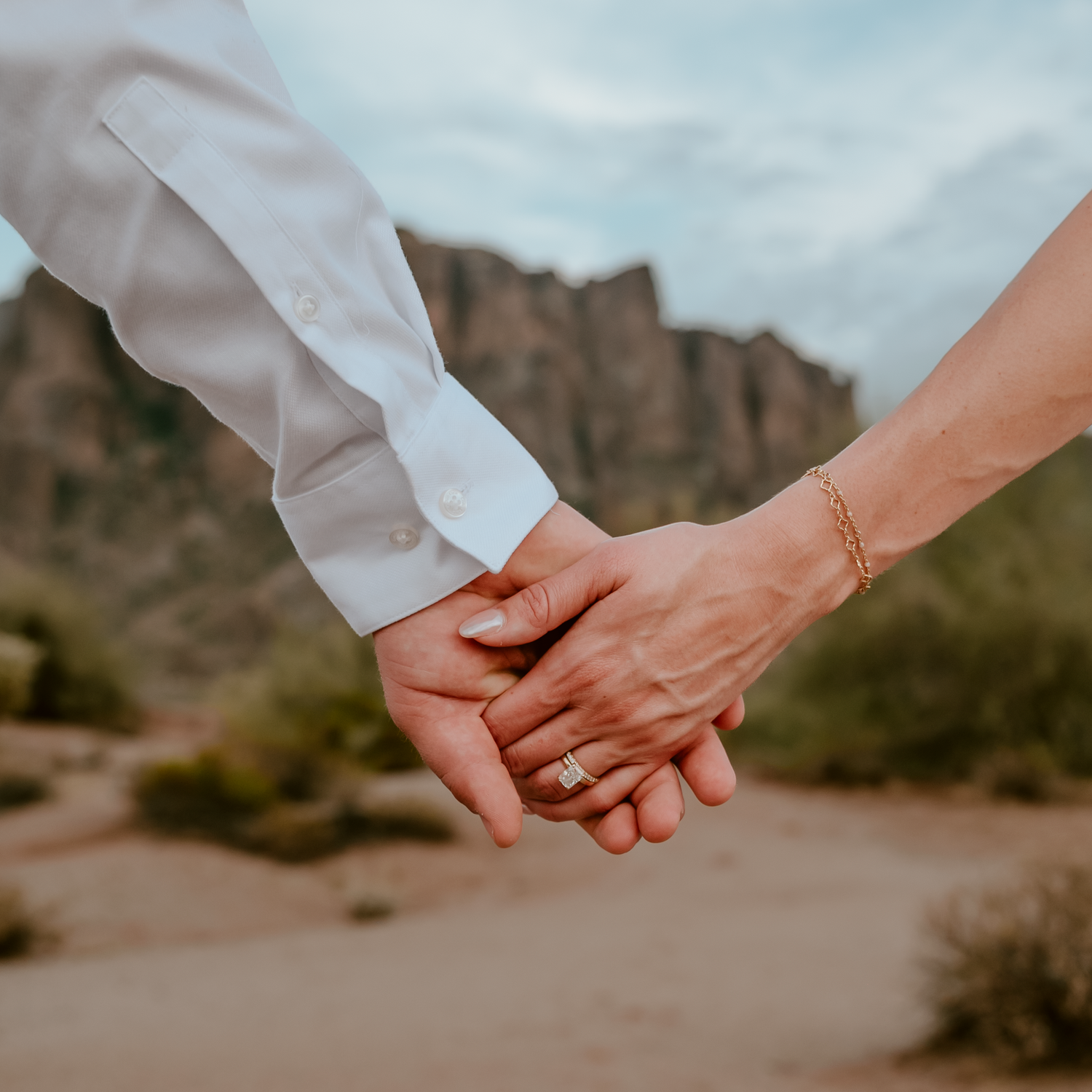 Couple holding hands in desert landscape, woman wearing gold wedding ring and bracelet, rocky mountains in background.