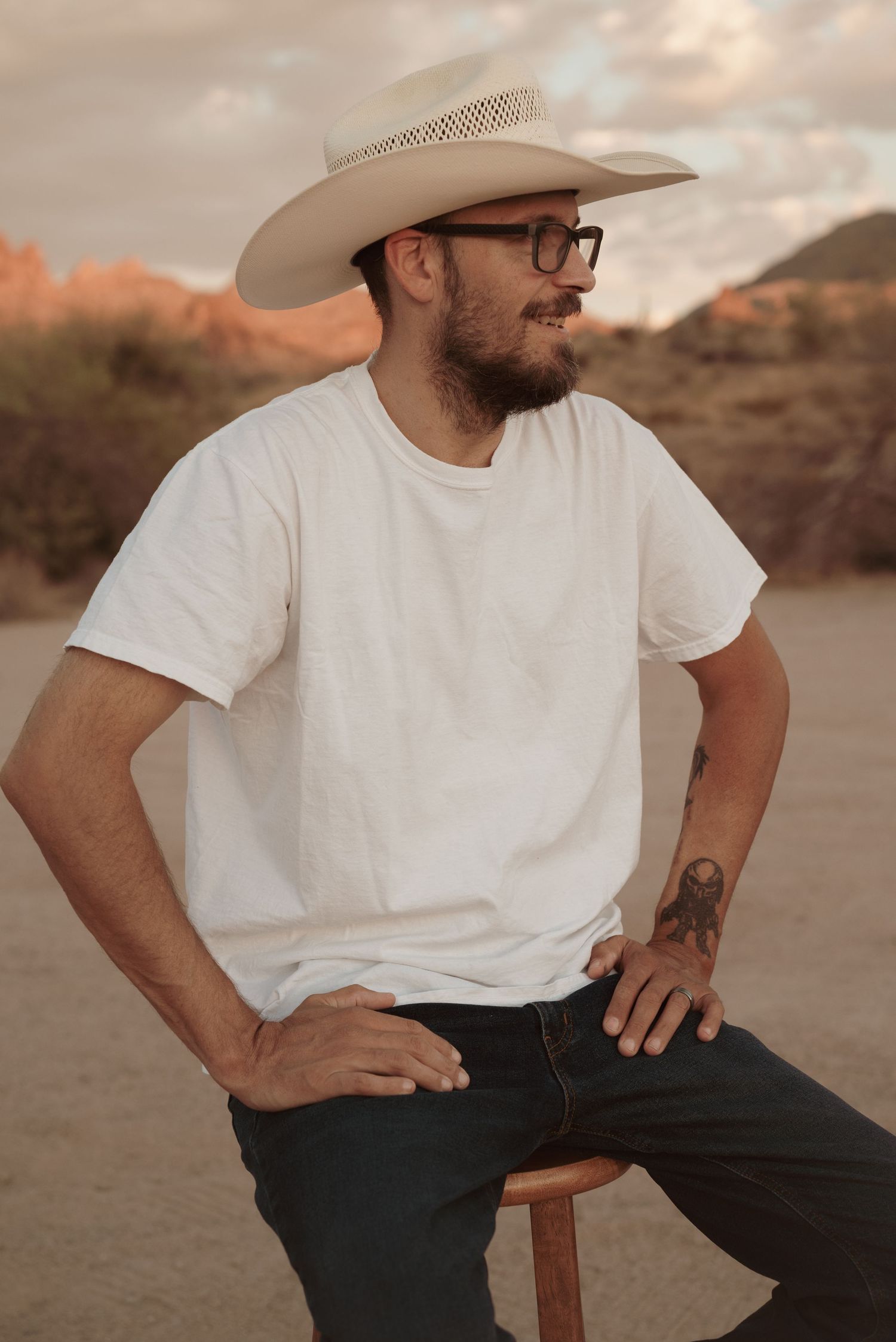 Bearded man in white cowboy hat, glasses, and white t-shirt sitting outdoors in desert landscape.