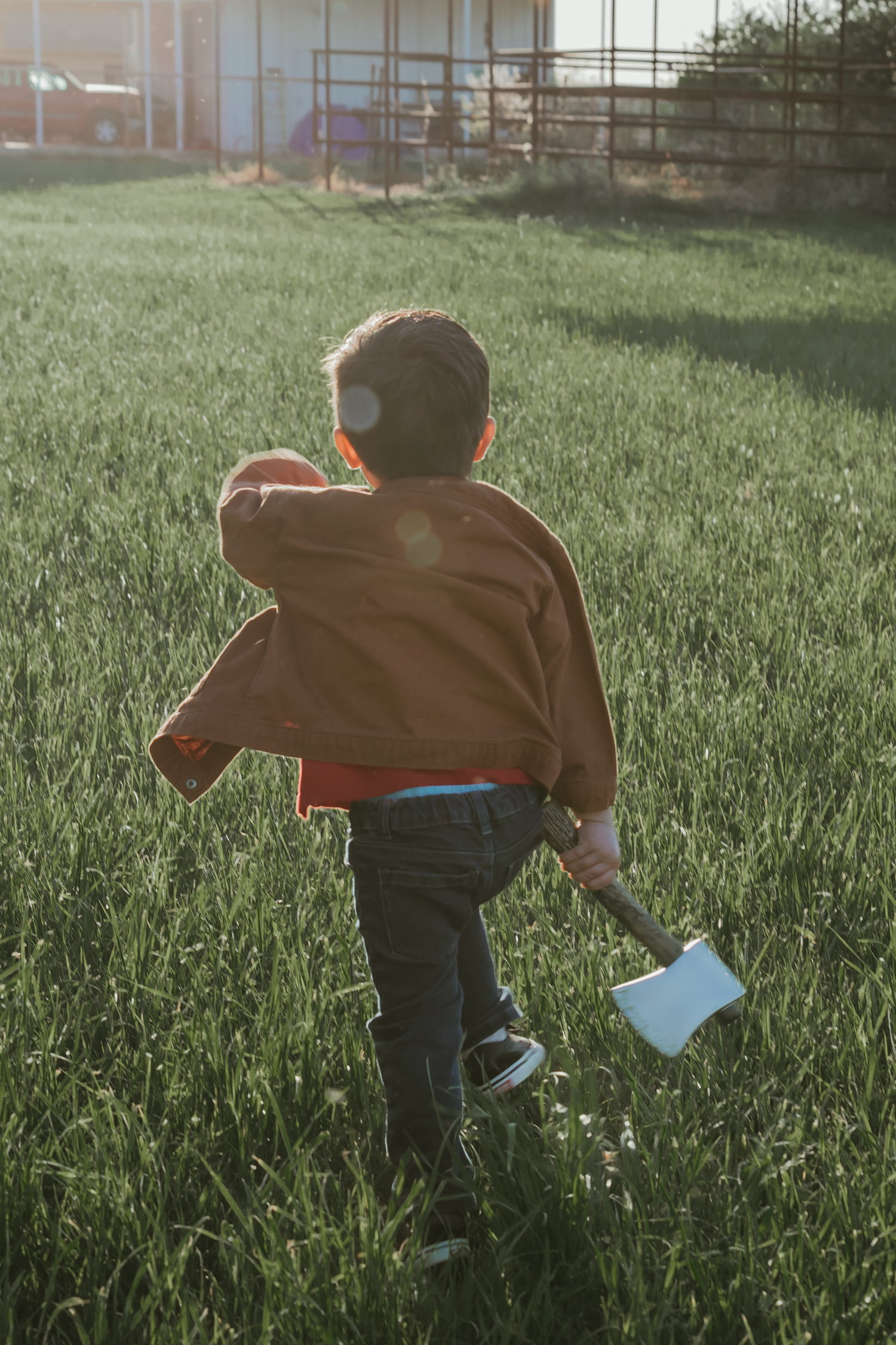 Young boy walking through green grass field carrying a toy axe, wearing brown jacket and jeans in golden sunlight.