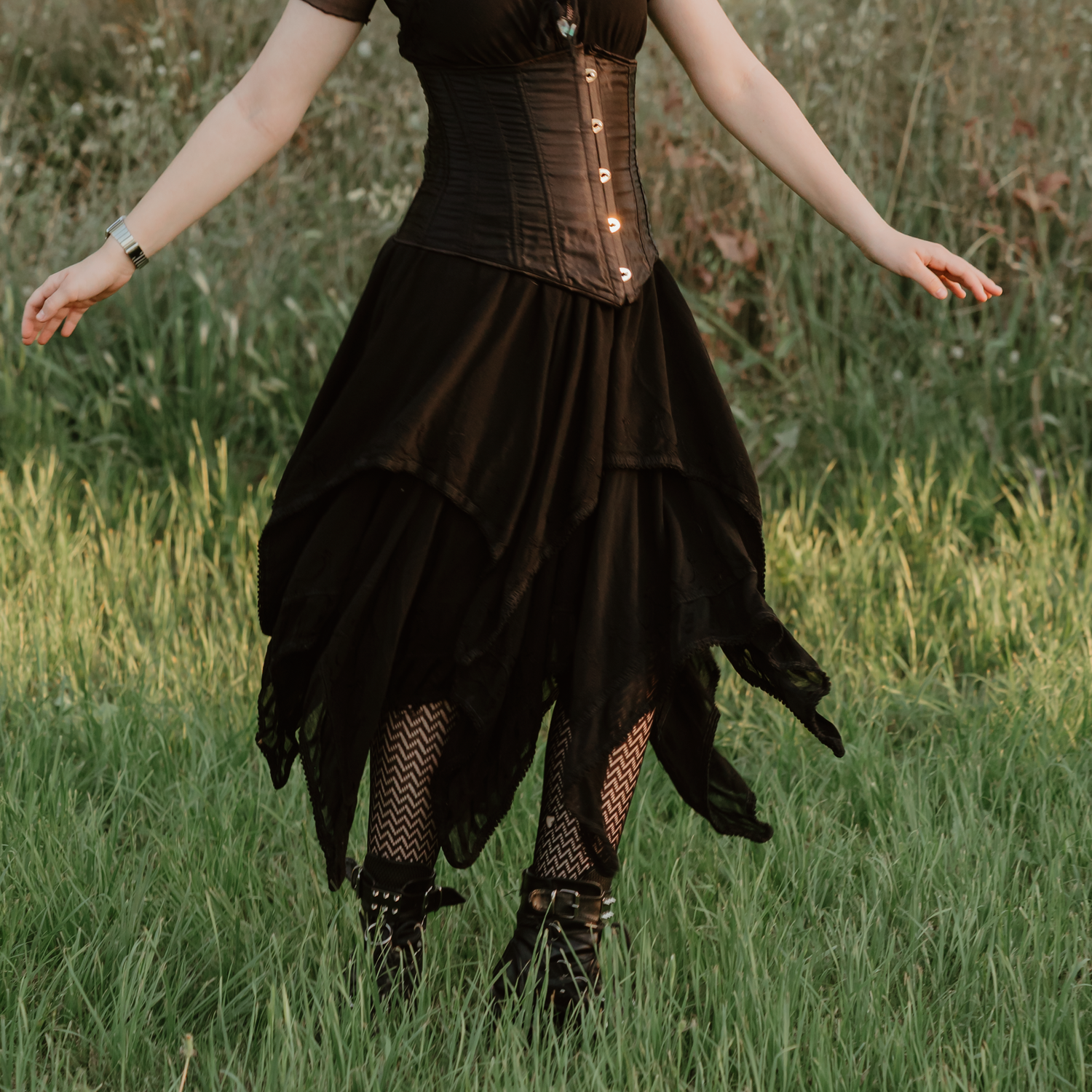 Woman in steampunk corset, black asymmetrical skirt, fishnet tights, and lace-up boots standing in a grassy field.