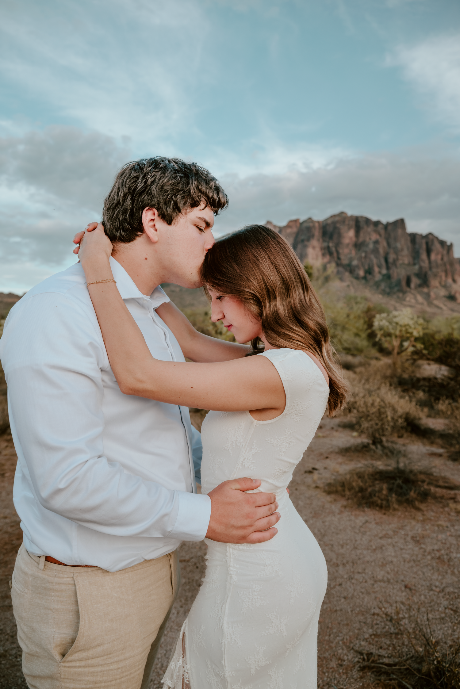 A couple embraces tenderly in a desert landscape with mountains and cacti, the man kissing the woman's forehead.