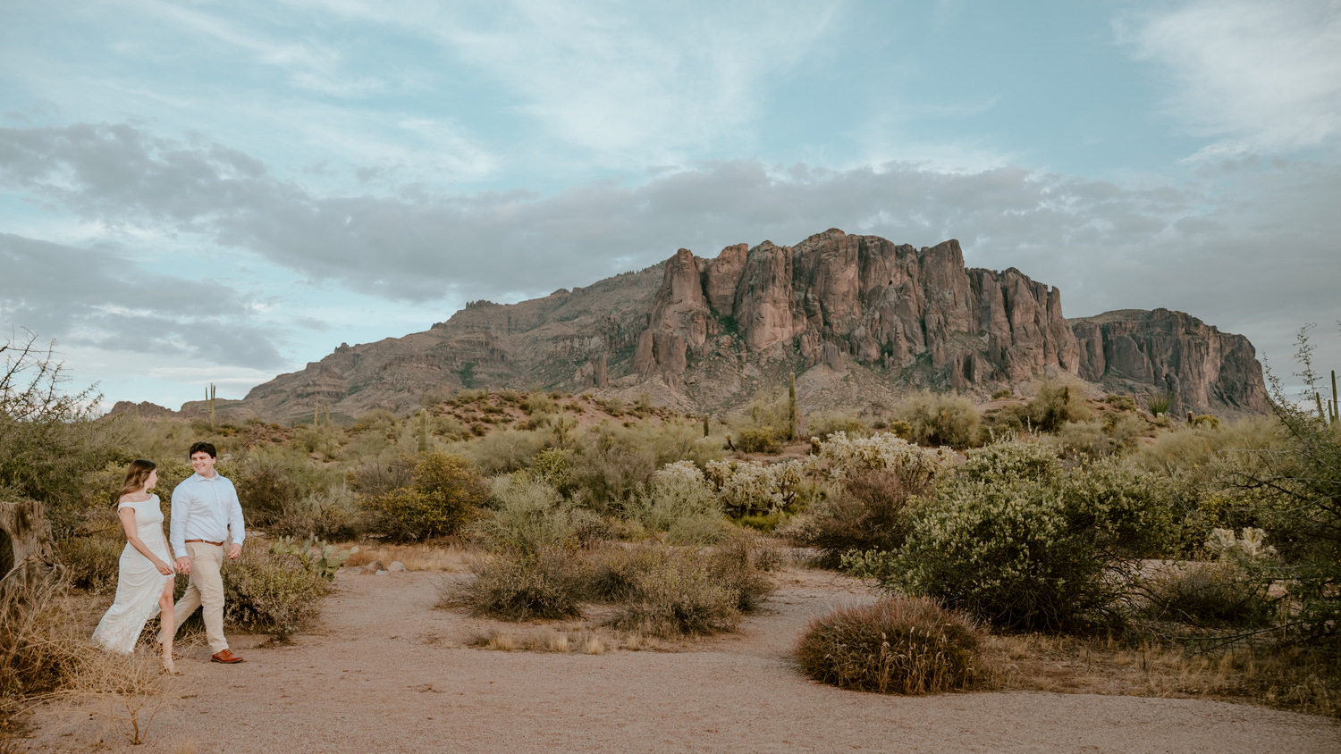 Couple walks hand-in-hand through Arizona desert with Superstition Mountains backdrop at dusk.