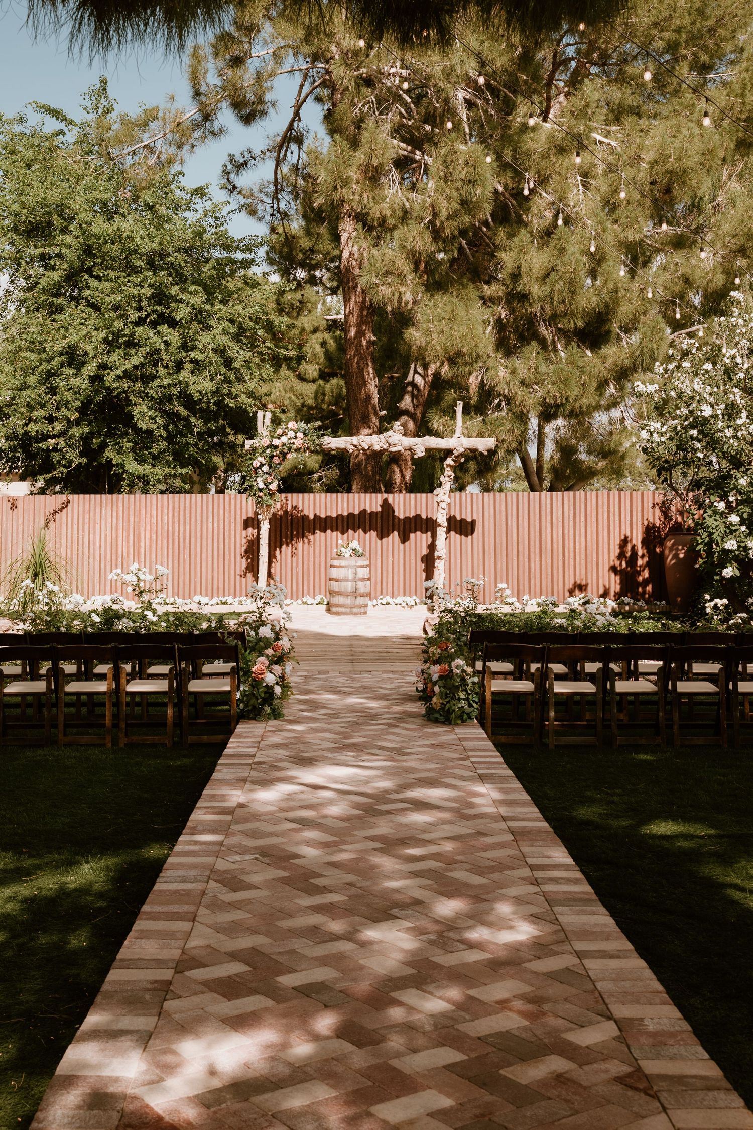 Brick paved pathway through garden with pink flowering trees and wooden fence.