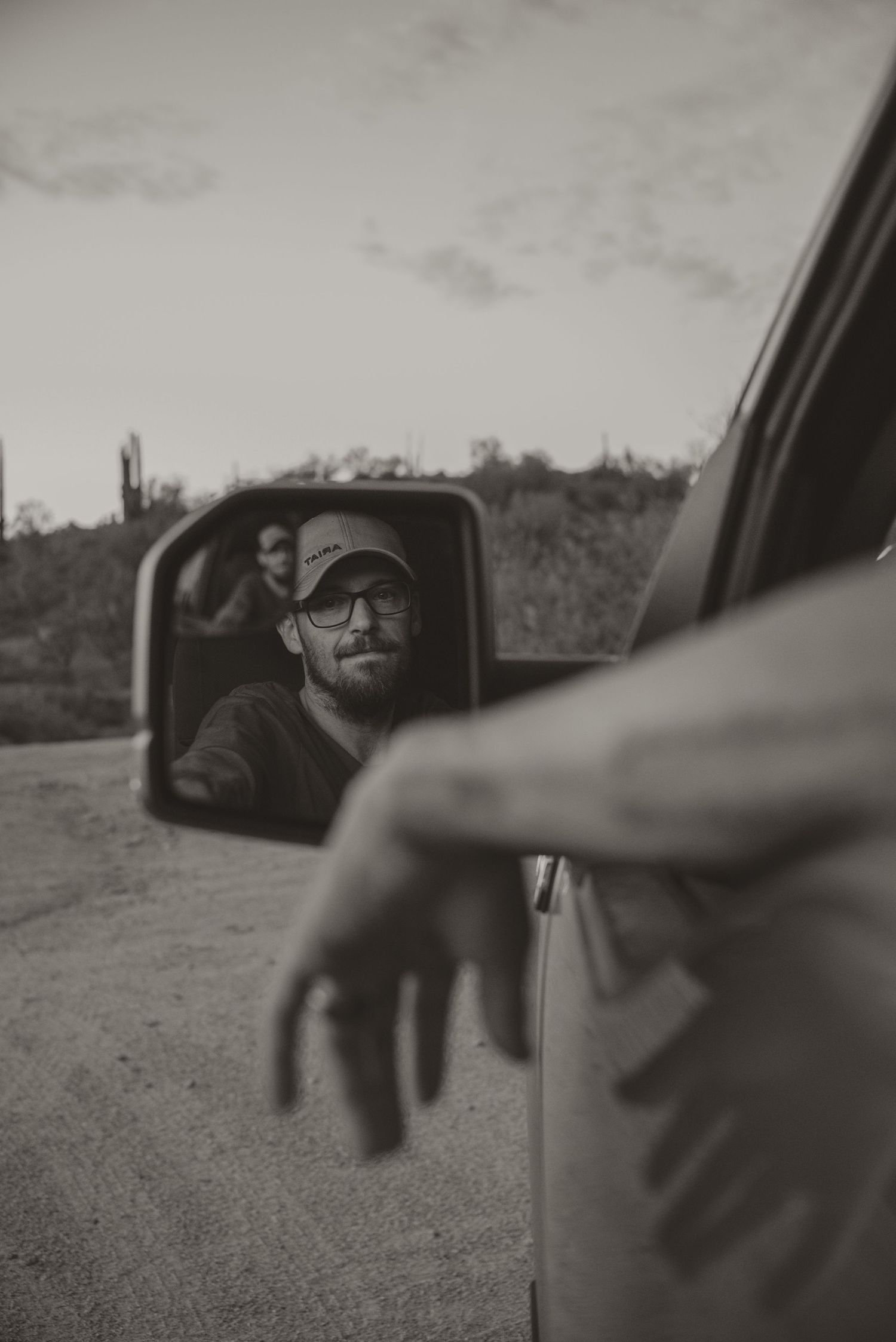 Man with glasses and cap reflected in car side mirror, arm hanging out window, desert landscape background.