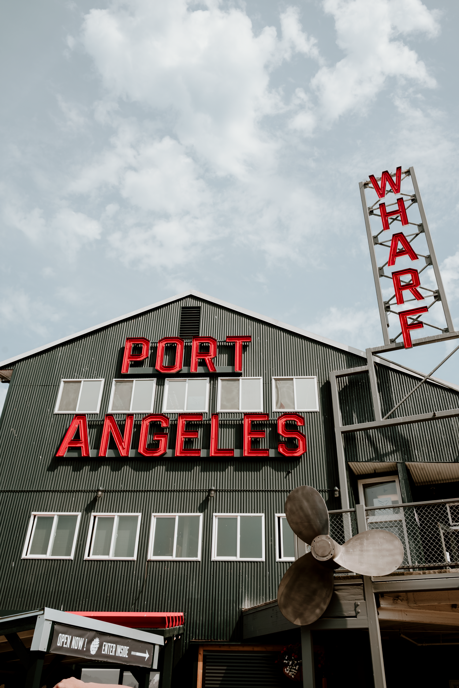 Red neon Port Angeles sign on a dark green wharf building with a ship propeller and Wharf sign against a cloudy sky.
