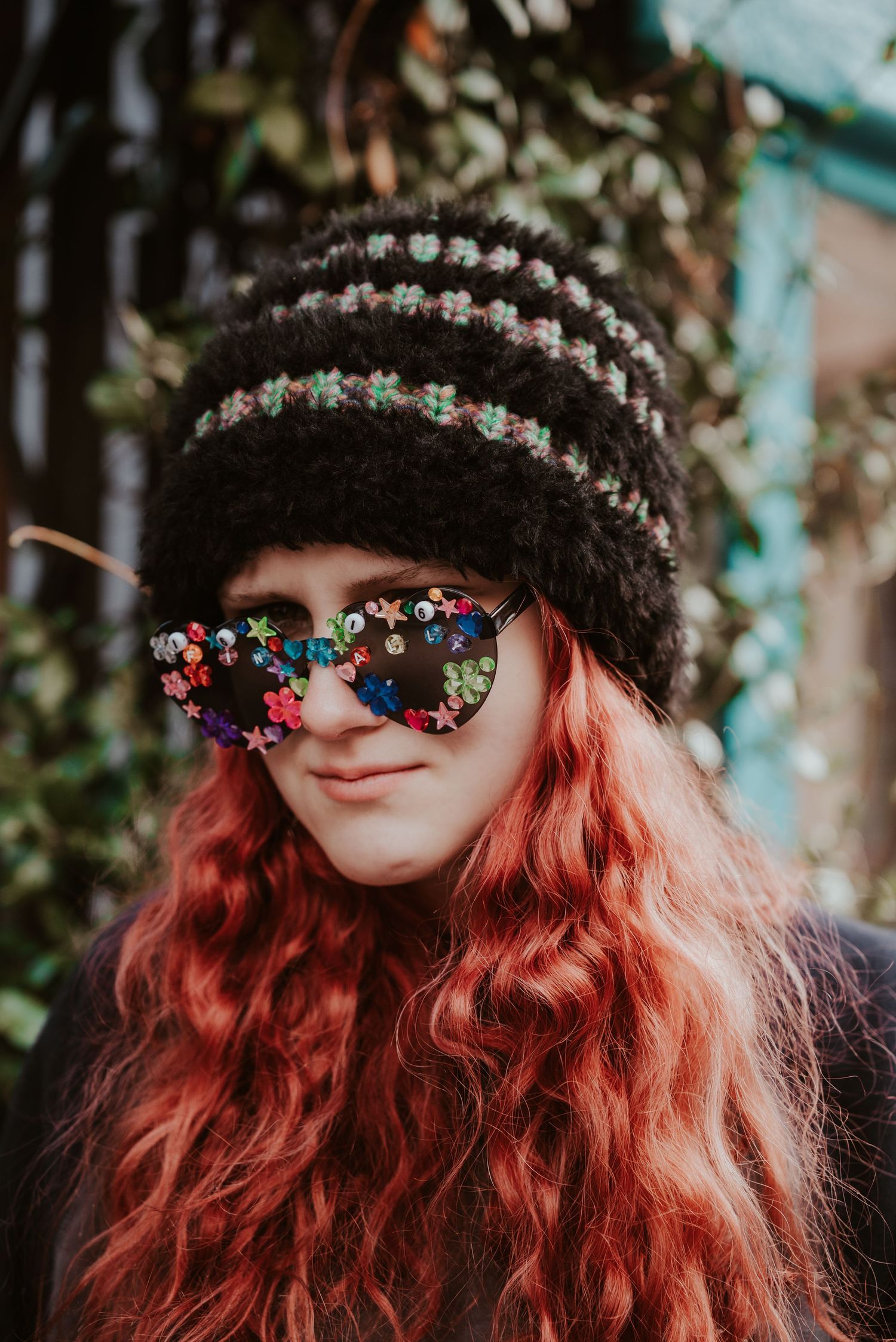 Young woman with red wavy hair wearing a striped knit beanie and decorative star-shaped novelty sunglasses outdoors.