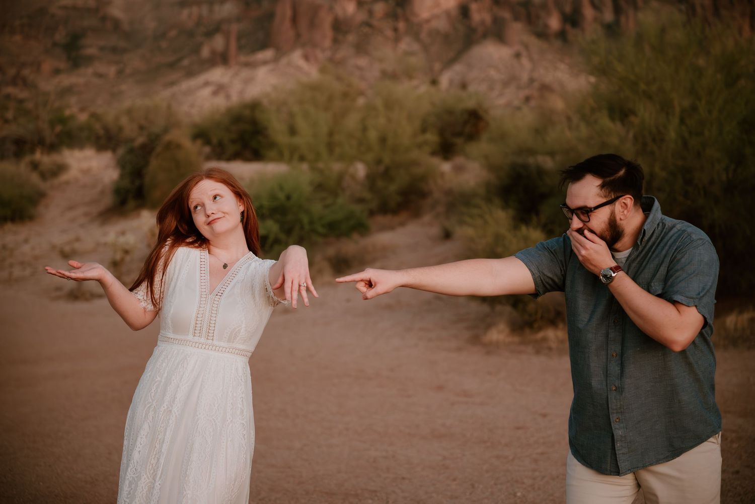 Couple laughing and playfully pointing at each other in a desert landscape with red rock formations in the background.