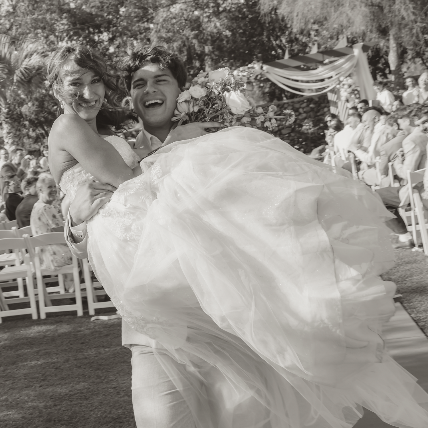 Black and white photo of a joyful bride being carried by groom at outdoor wedding ceremony.