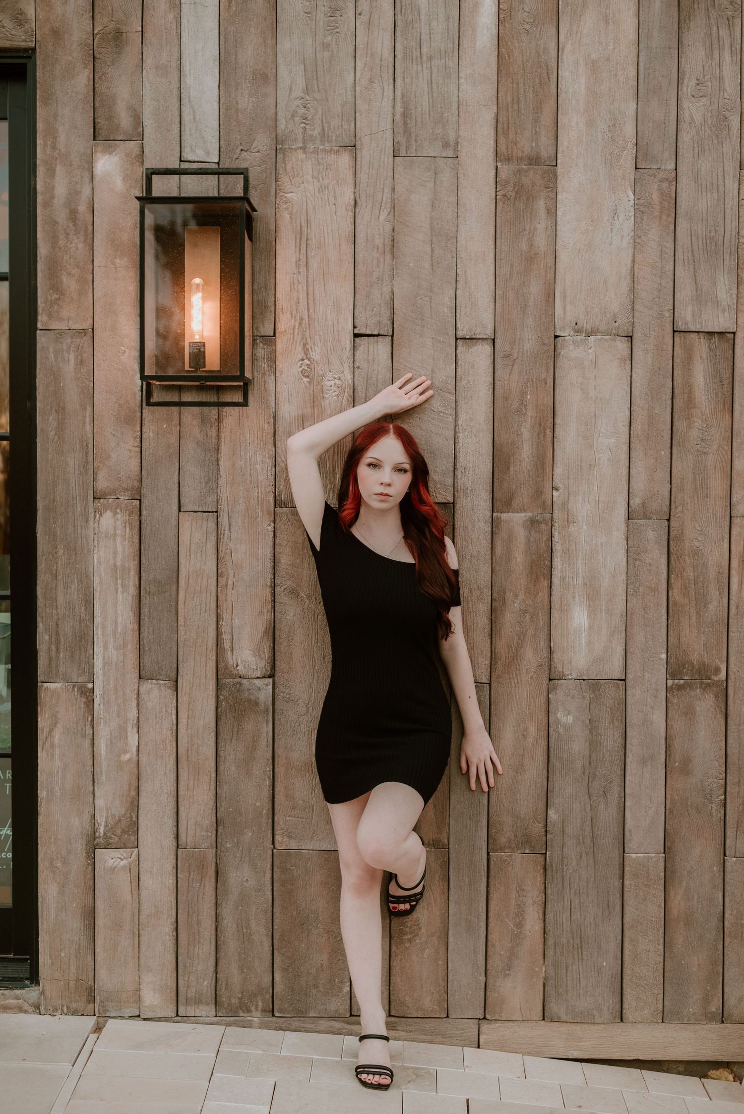 Fashion pose in fitted black dress against wooden wall with vintage lantern.