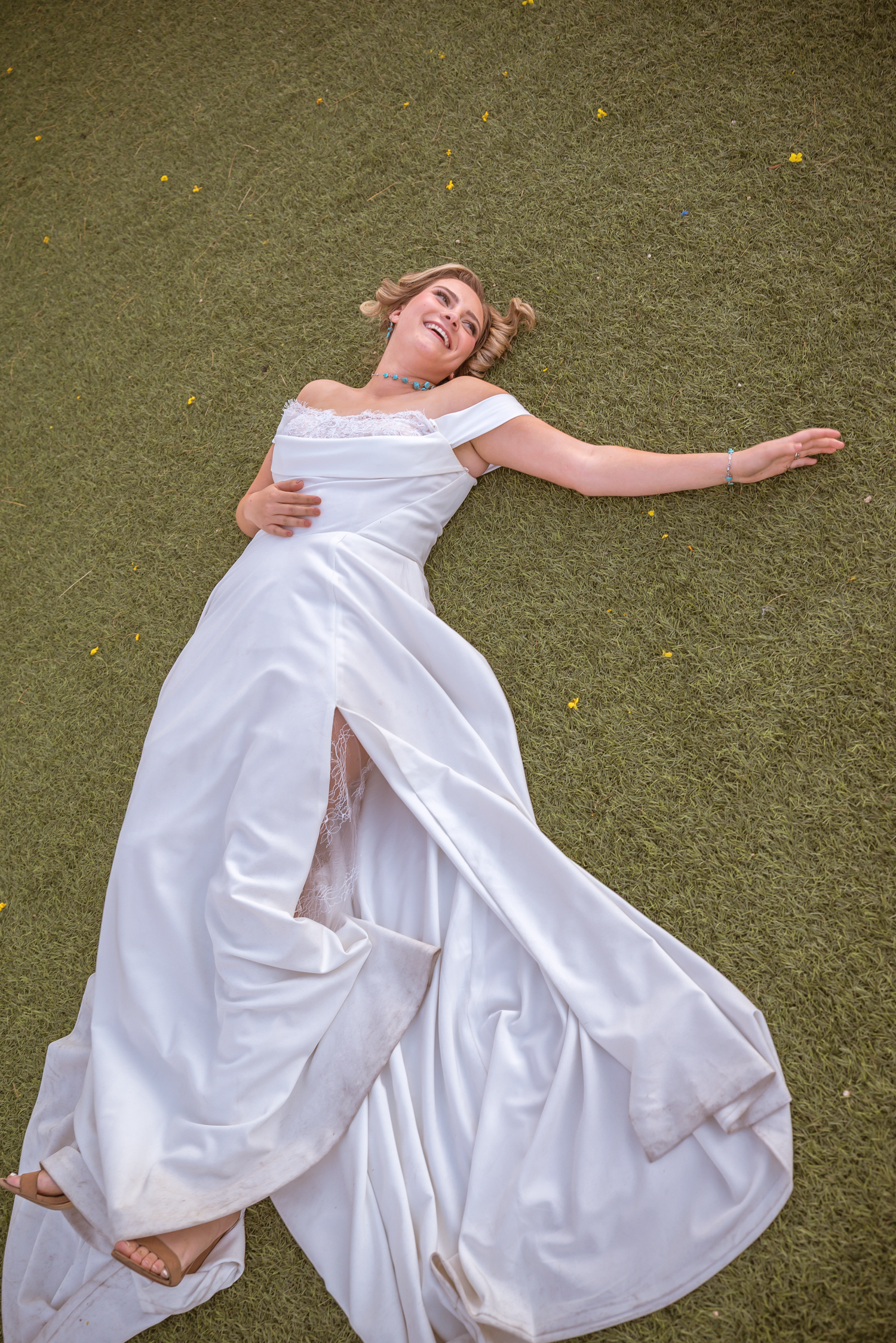 Bride in white off-shoulder gown lying on green grass, laughing joyfully with arms spread wide.