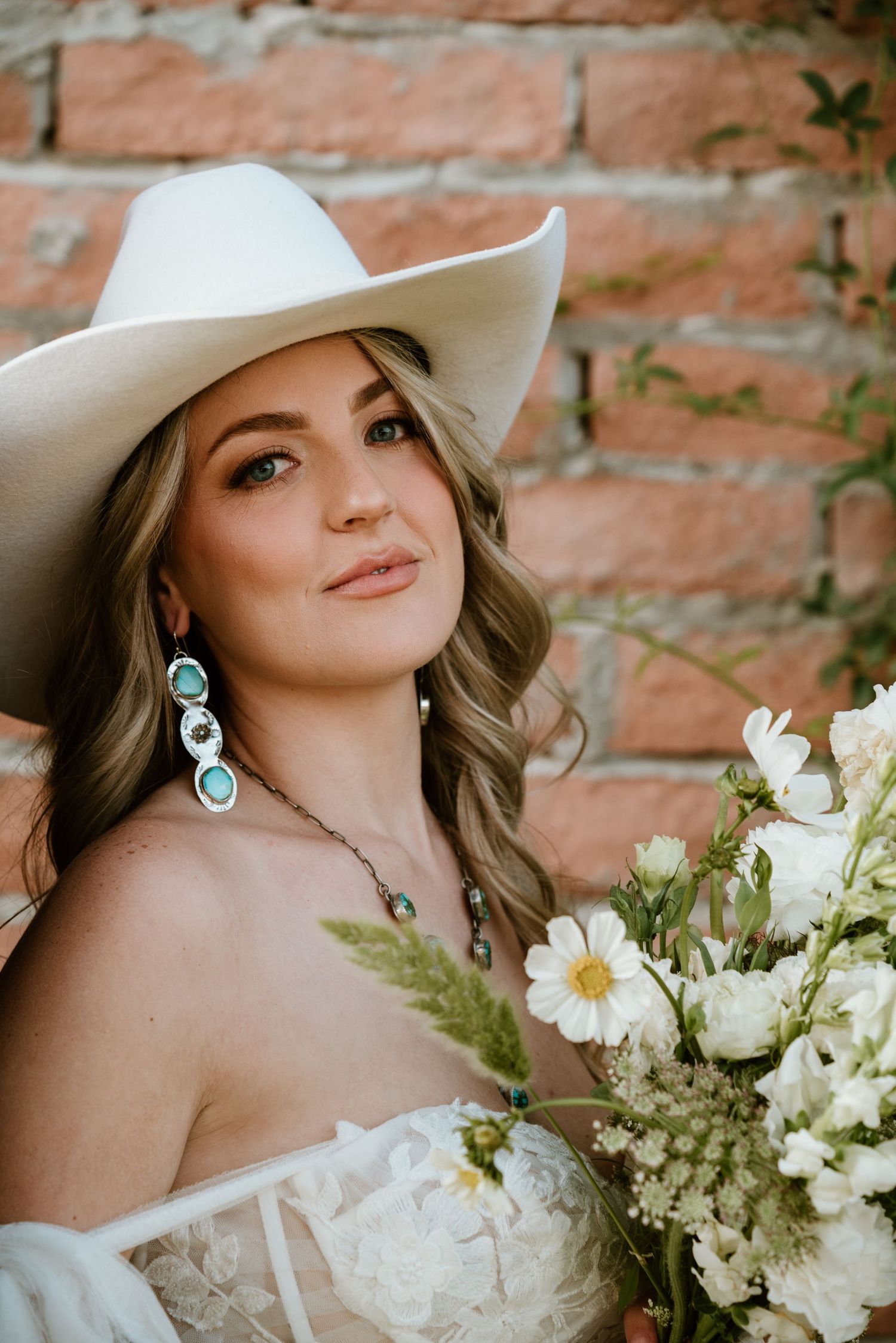 Bridal portrait with white cowboy hat and turquoise jewelry against brick wall.