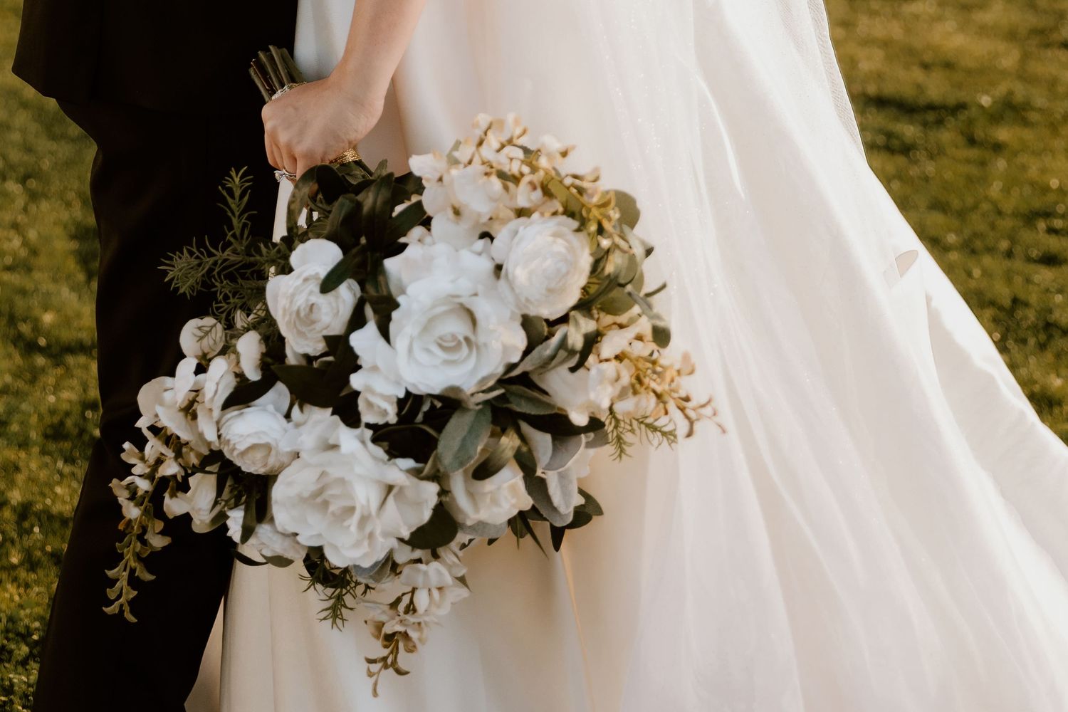 An elegant white wedding bouquet with roses and delicate greenery held against a white dress in natural sunlight.