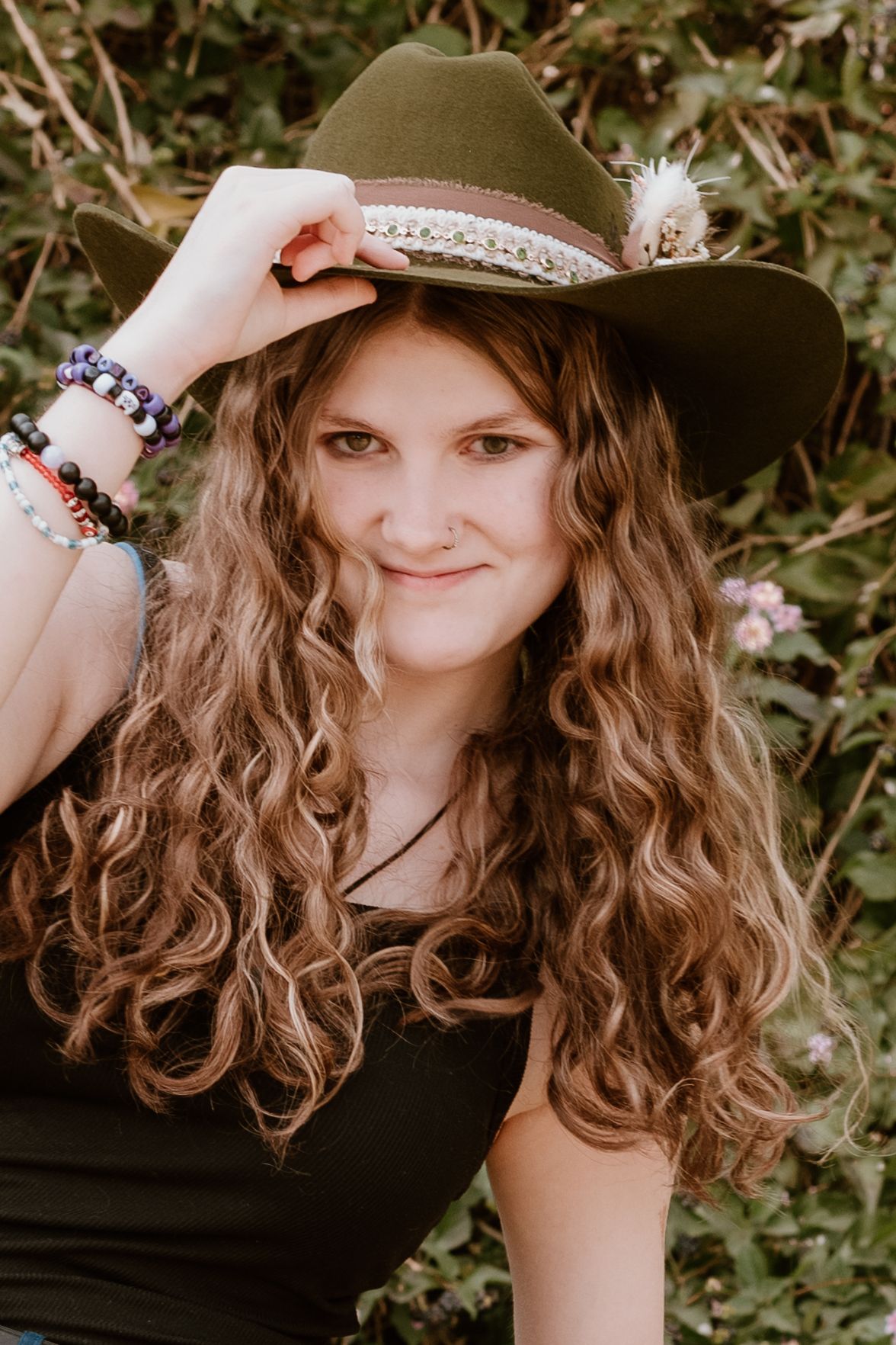 Natural outdoor photo showing person wearing white hat with curly hair against green foliage.