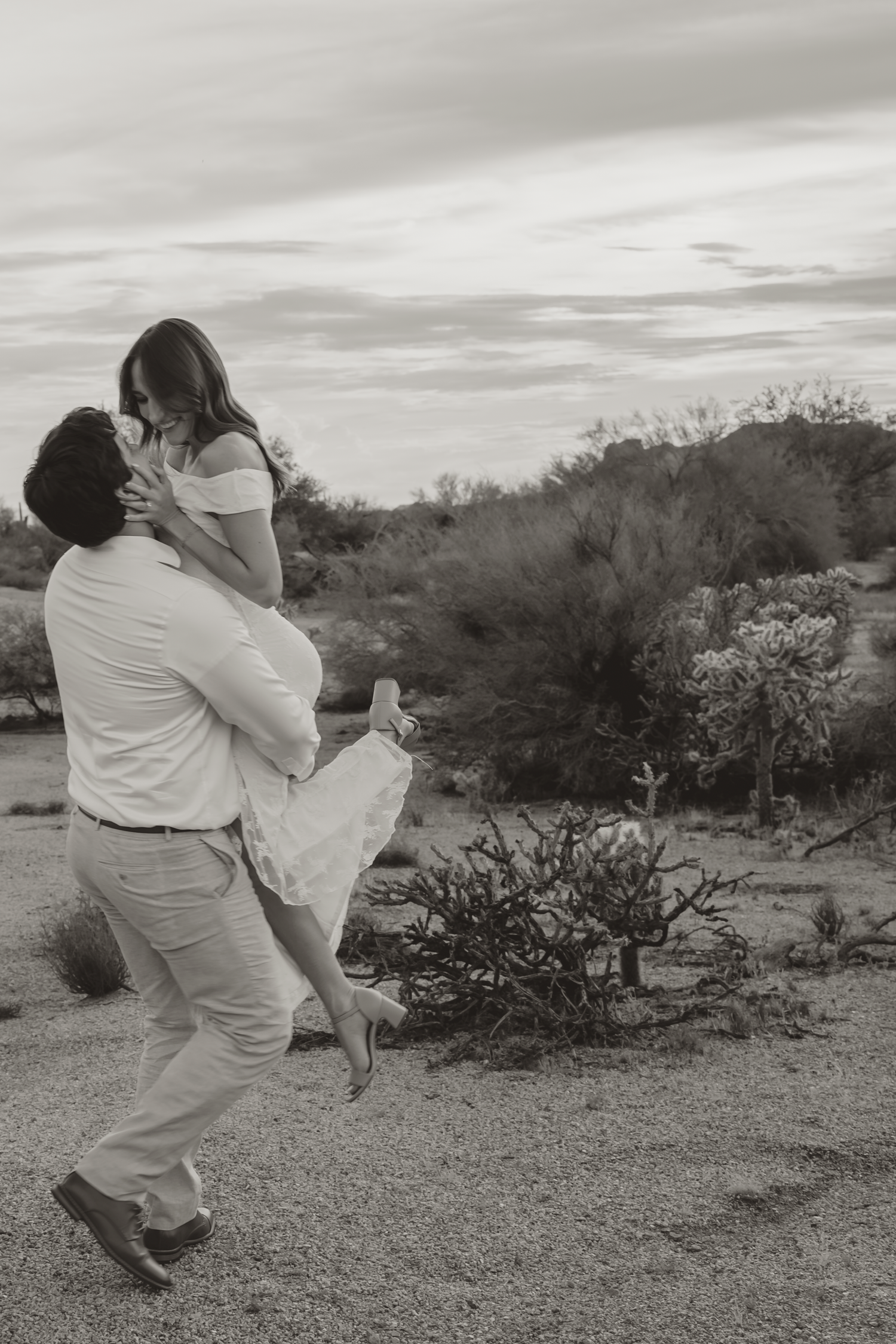 A man lifts and embraces a woman in a romantic desert landscape, captured in black and white photography.