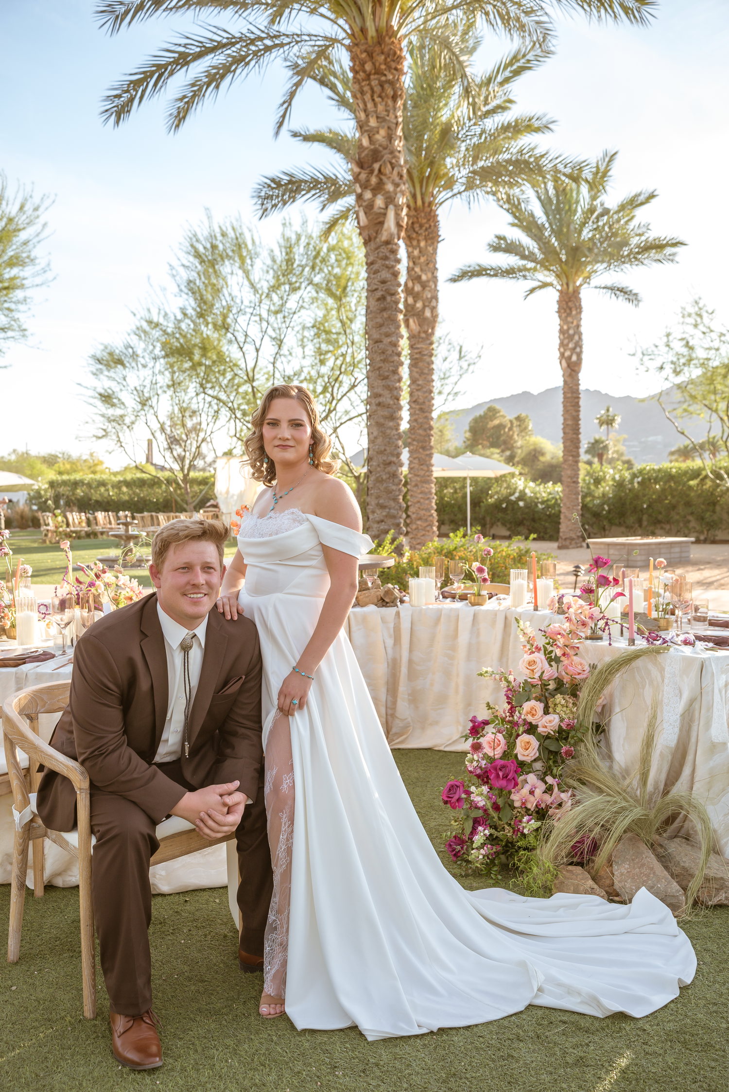 Bride in white gown and groom in brown suit pose outdoors at elegant wedding reception with palm trees and floral arrangements.