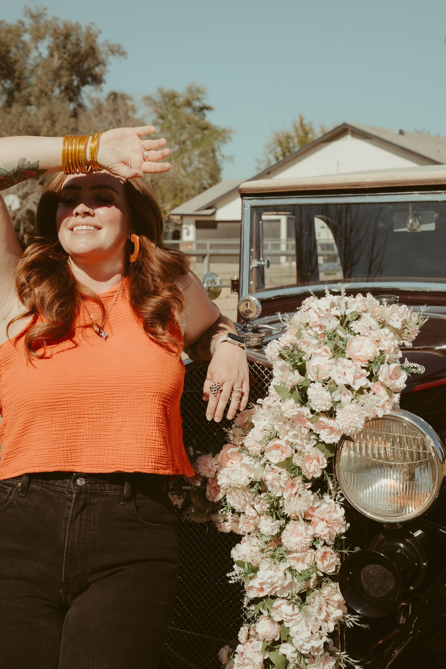 Woman in orange top poses by vintage car adorned with pink floral garland, wearing gold bangles and orange earrings.