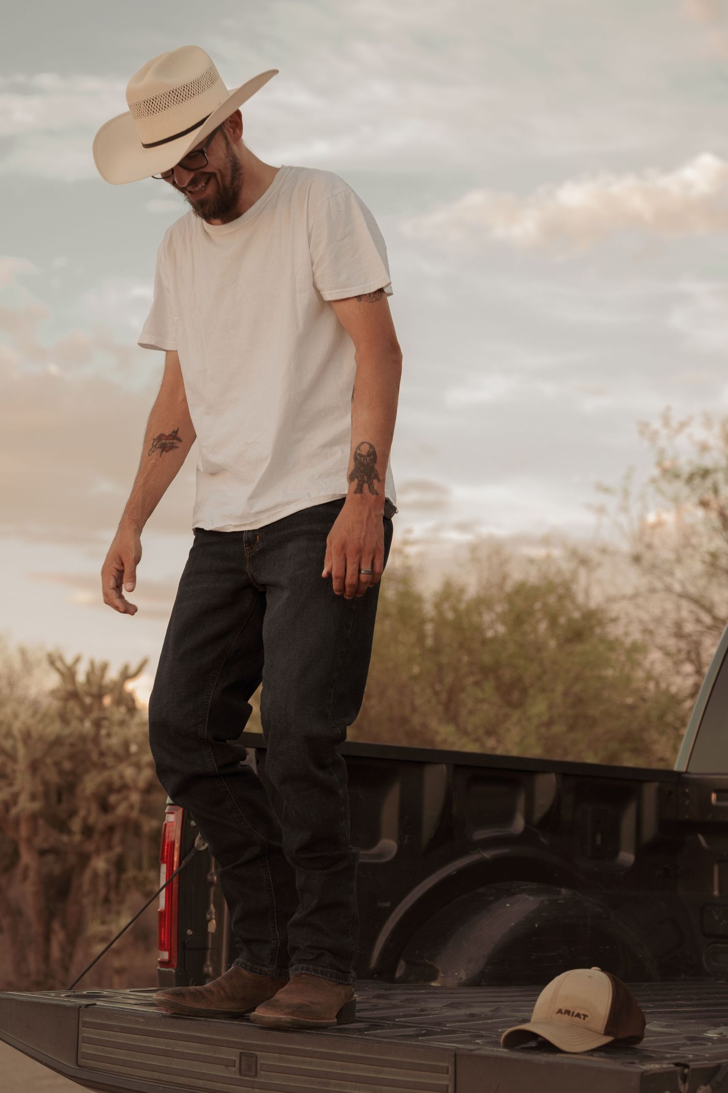 Tattooed man in cowboy hat and white tee stands on truck bed in desert setting at dusk.