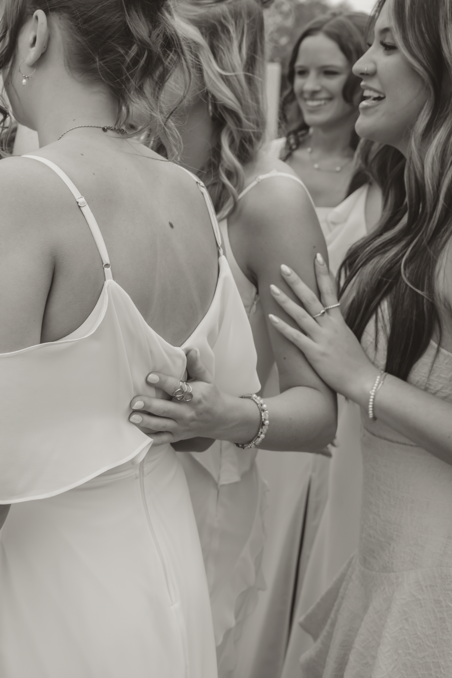 Black and white photo of bridesmaids helping bride with wedding dress buttons, smiling and laughing together.