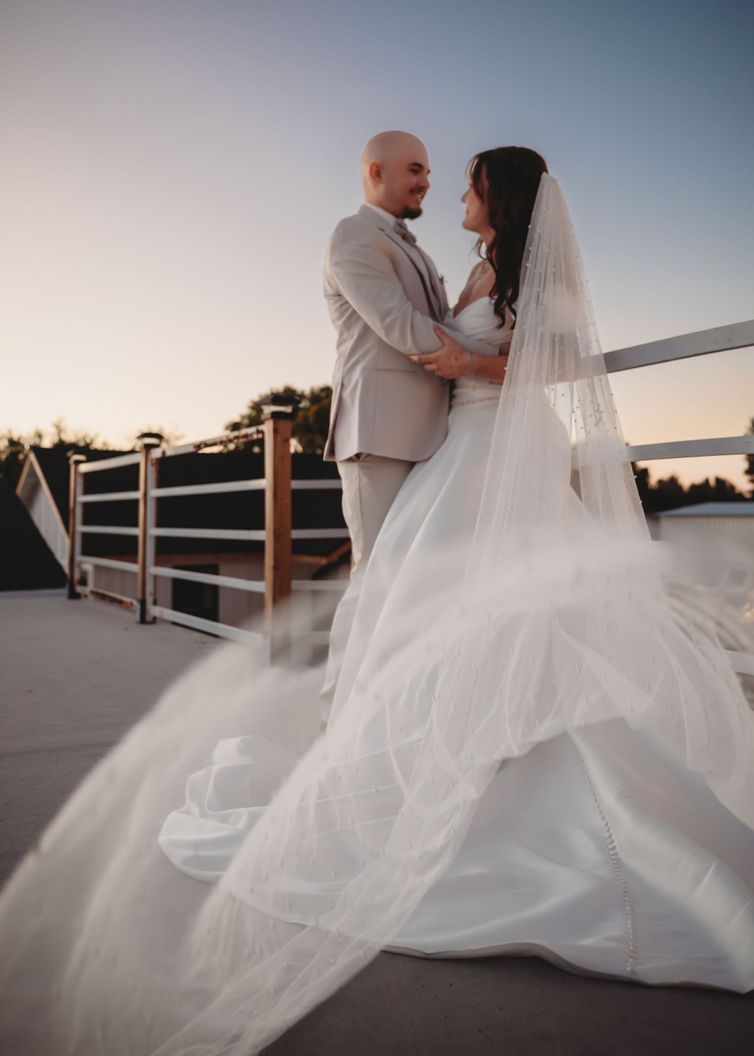Bride in flowing white gown and veil stands with groom in tan suit on outdoor deck at sunset.