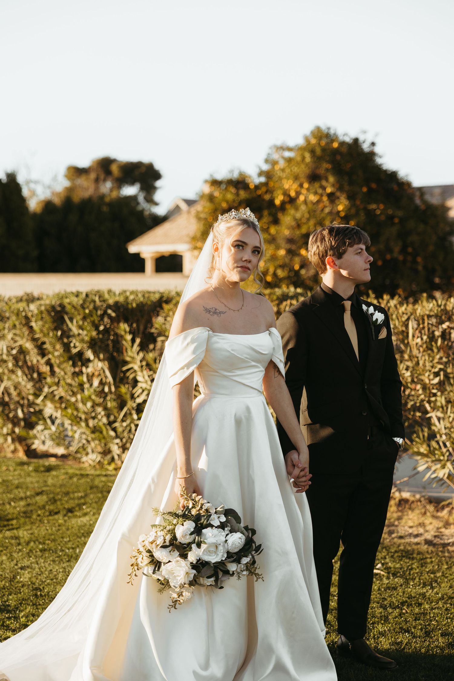 A couple in wedding attire walking through a sunlit garden.