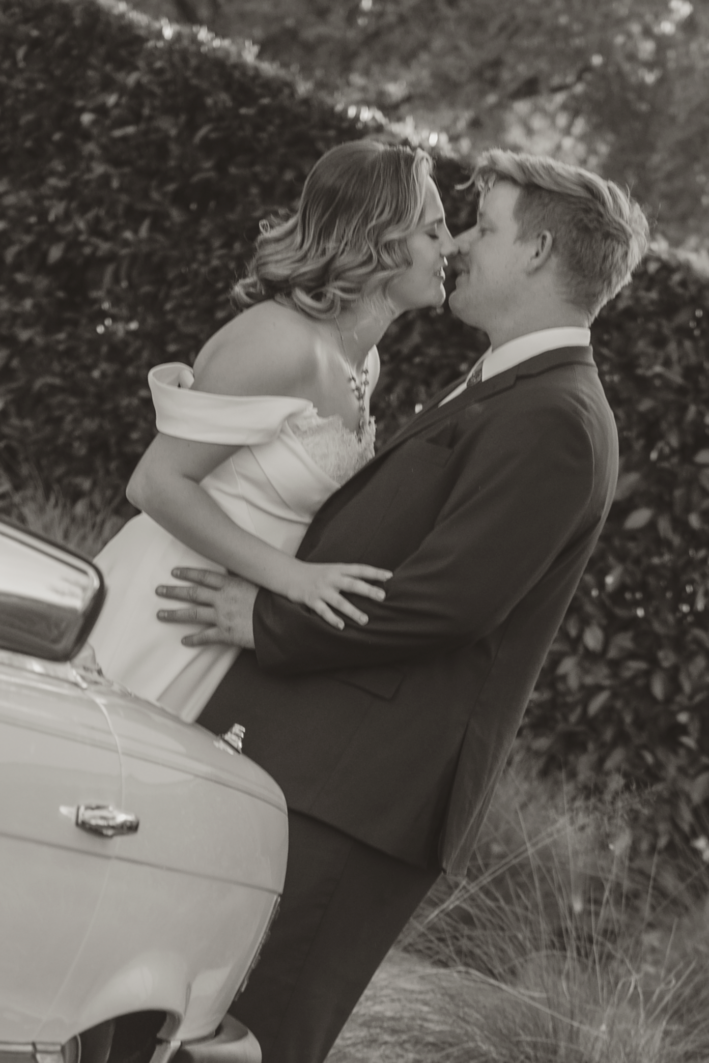 Black and white photo of a bride and groom sharing a romantic kiss beside a vintage white car outdoors.