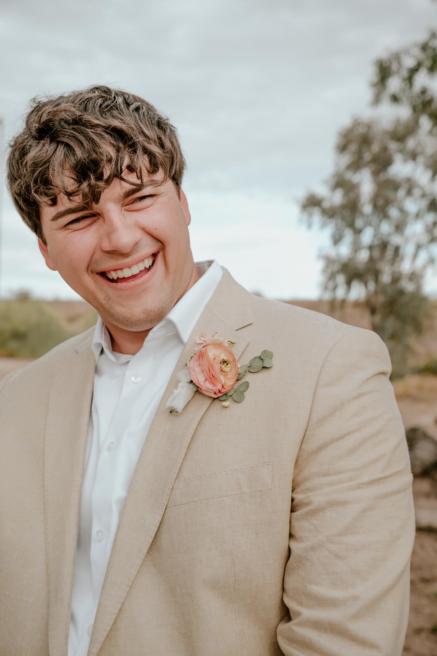 Smiling groom in a tan linen suit with a pink rose boutonniere at an outdoor desert wedding.