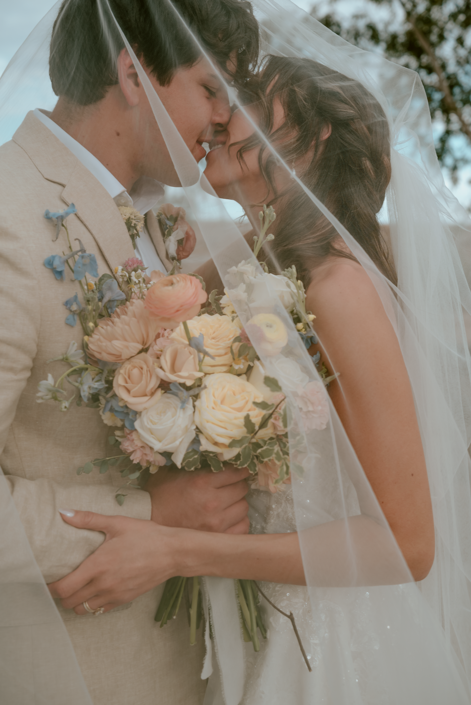 Bride and groom share a romantic moment under a veil, holding a pastel bouquet of roses and wildflowers.