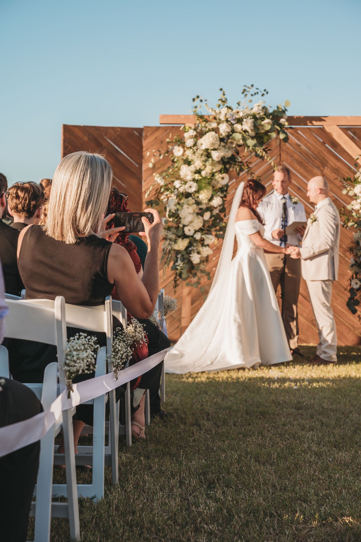 Outdoor wedding ceremony at sunset with floral arrangements and white chairs set up on a grassy lawn.