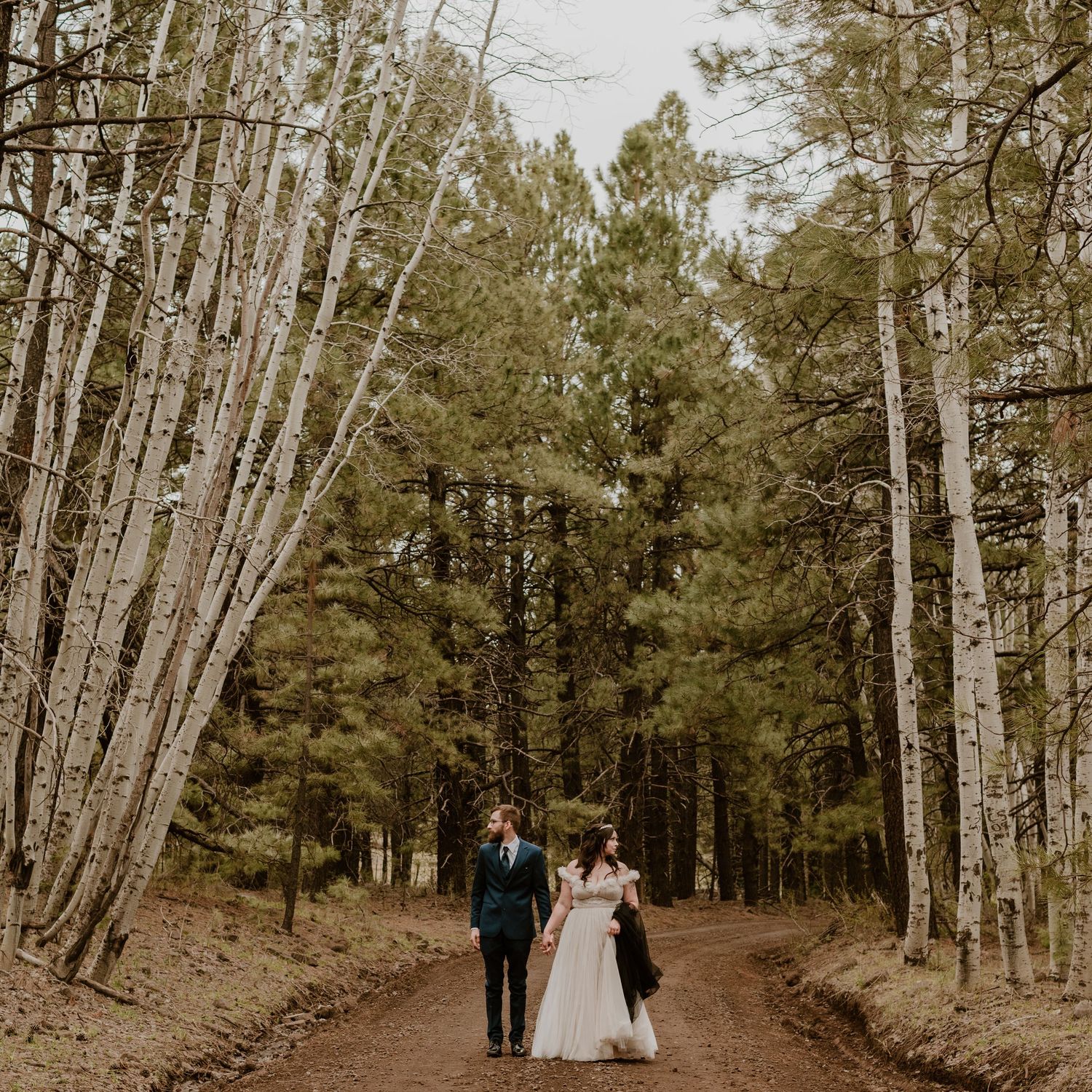 A woodland wedding portrait showing a couple walking down a forest path lined with tall trees.